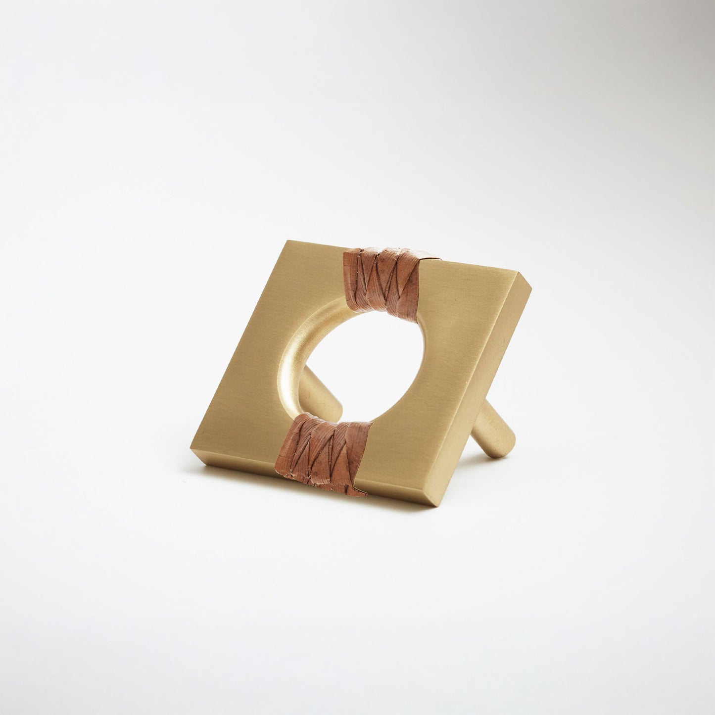 A satin brass metal geometric cabinet pull with a square frame and circular center, wrapped with brown leather at the corners, shown on a white background.