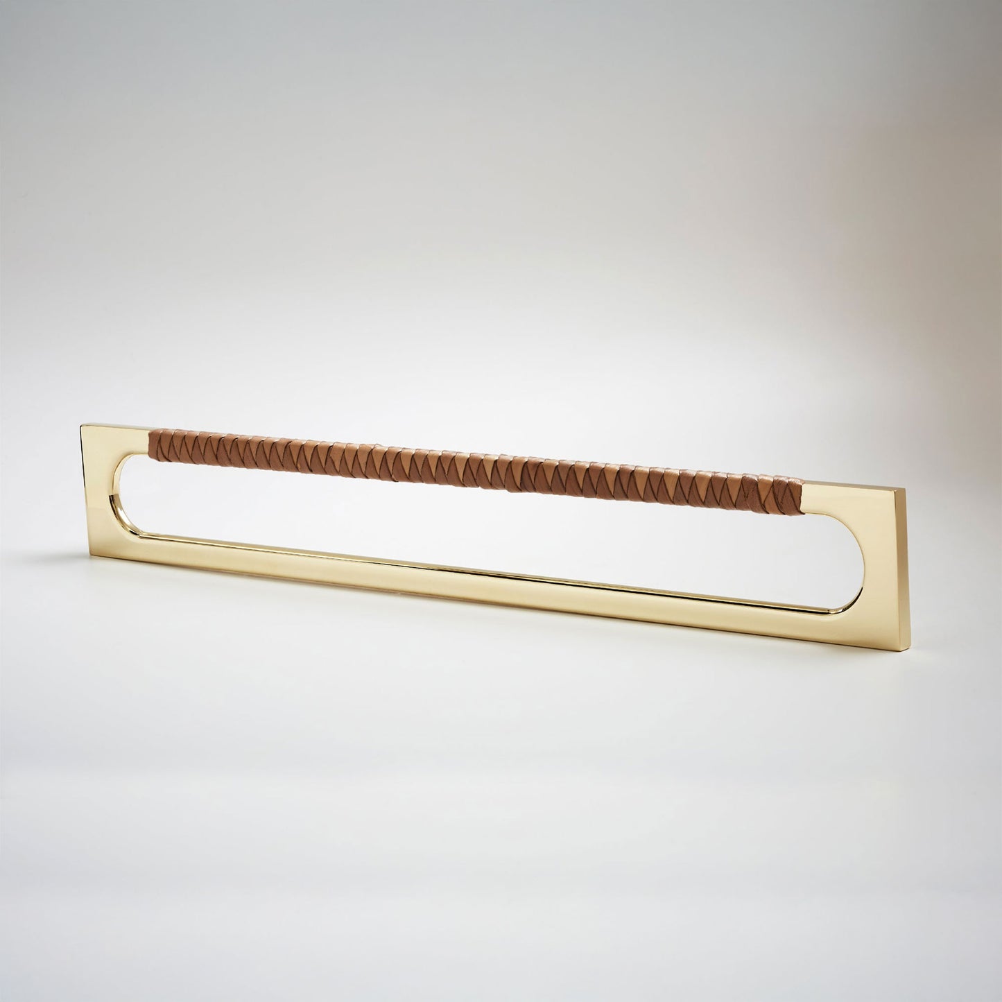 A modern long rectangular cabinet pull handle with a natural brass metal frame with an oval shaped-cutout and a brown leather woven grip, shown on a white background.