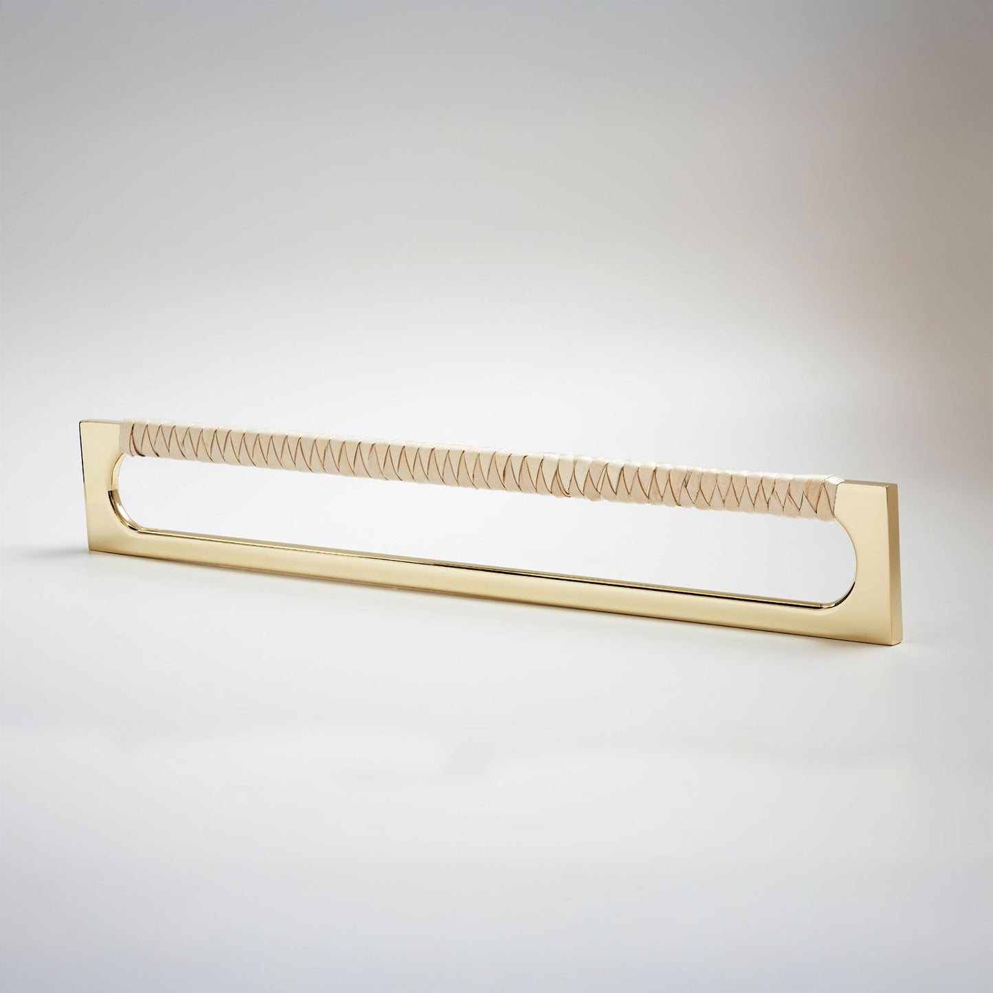 A modern long rectangular cabinet pull handle with a polished brass metal frame with an oval shaped-cutout and a light tan leather woven grip, shown on a white background.