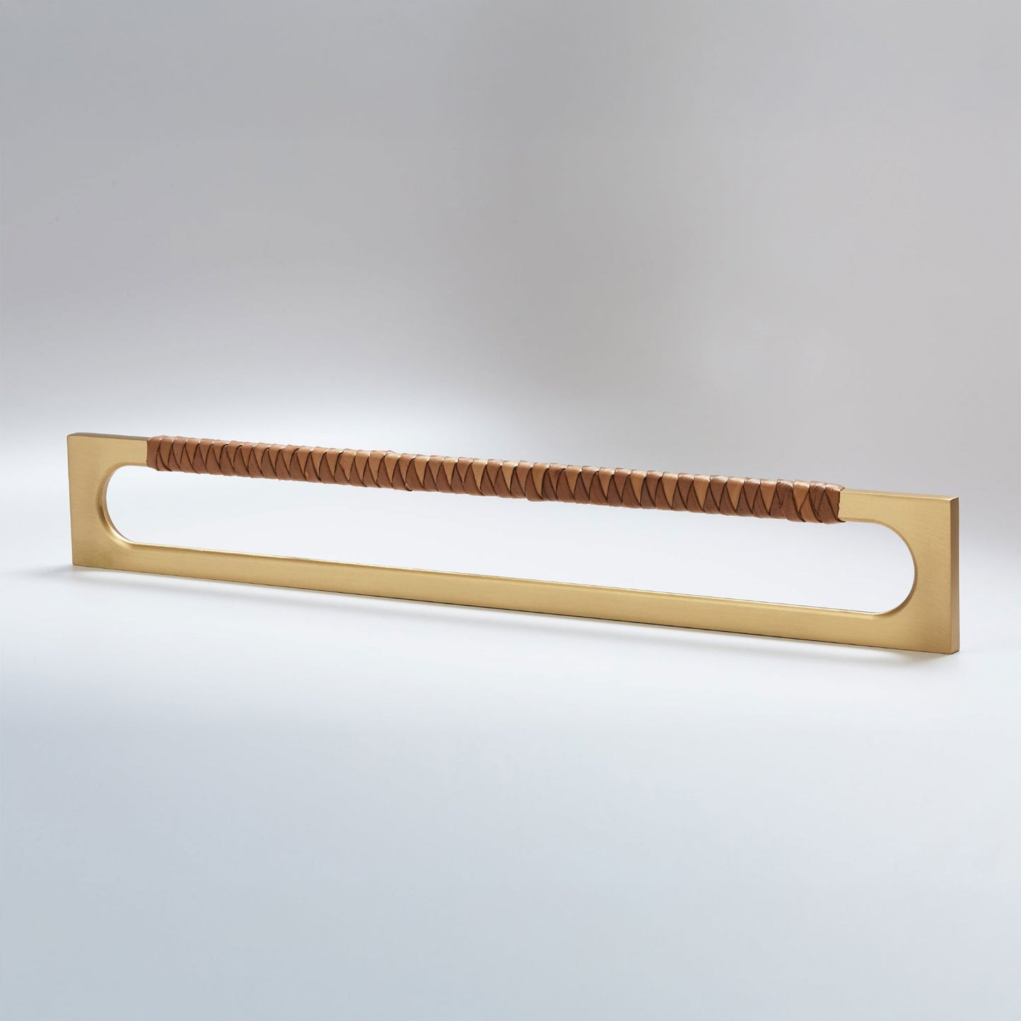 A modern long rectangular cabinet pull handle with a satin brass metal frame with an oval shaped-cutout and a brown leather woven grip, shown on a white background.