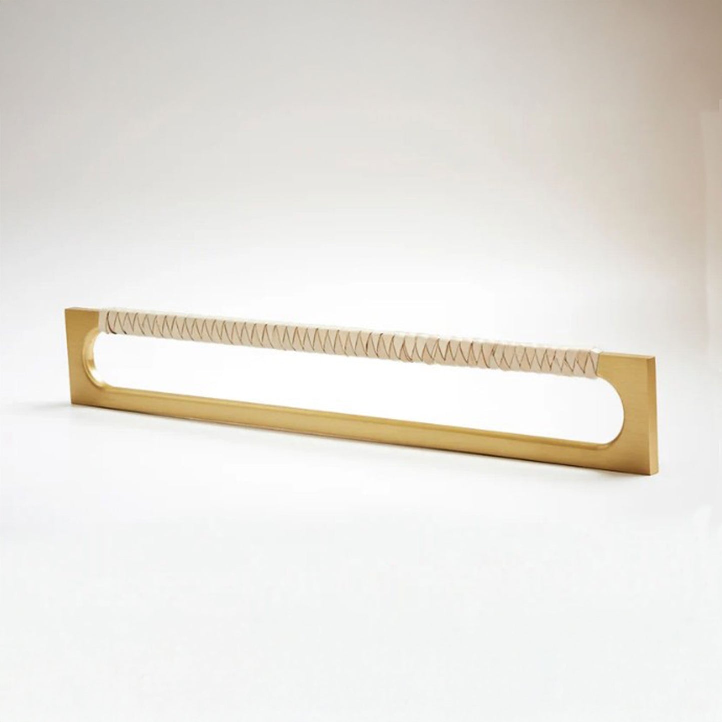 A modern long rectangular cabinet pull handle with a satin brass metal frame with an oval shaped-cutout and a light tan leather woven grip, shown on a white background.