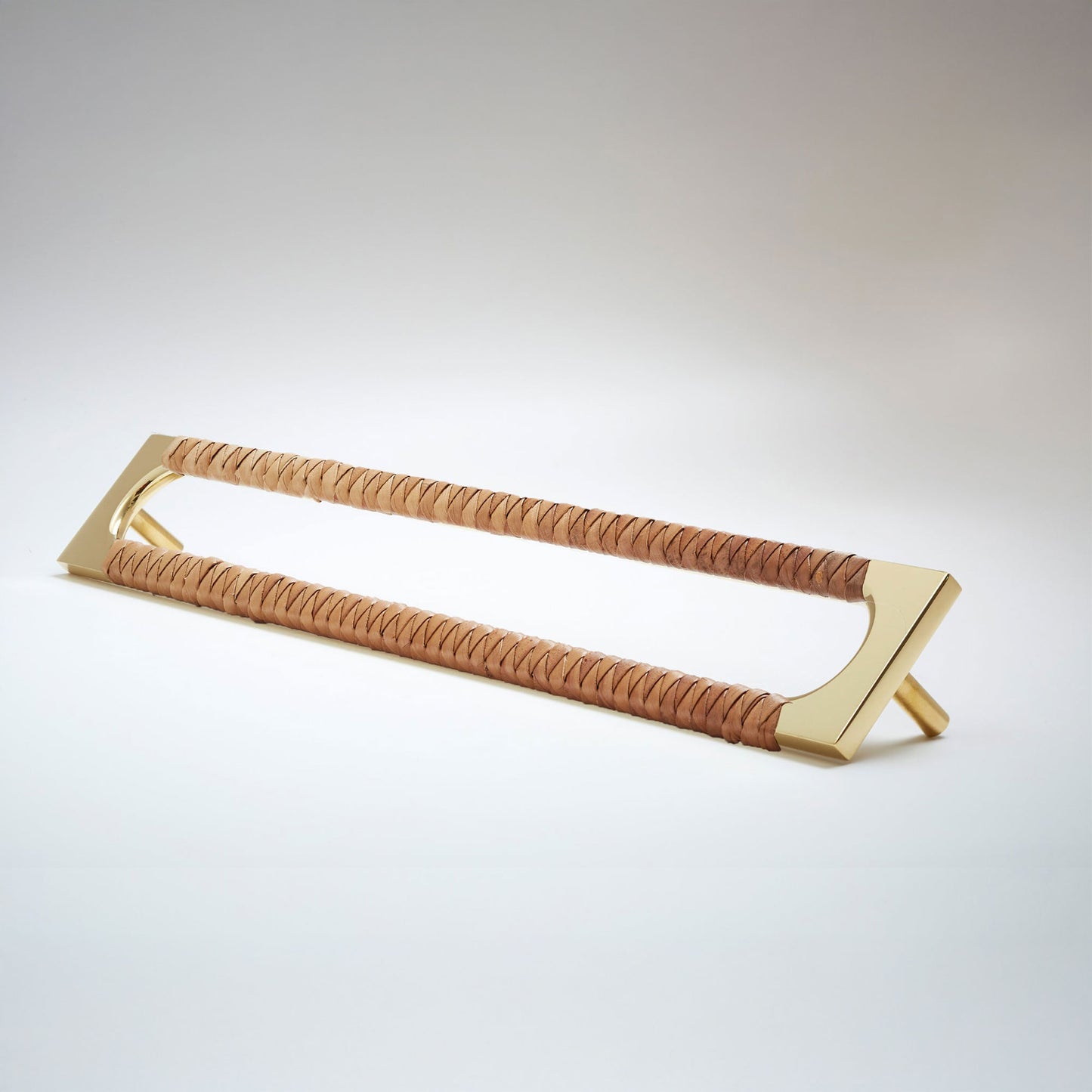 A modern long rectangular cabinet pull handle with a polished natural brass metal frame with an oval shaped-cutout and a brown leather woven grip, shown on a white background.