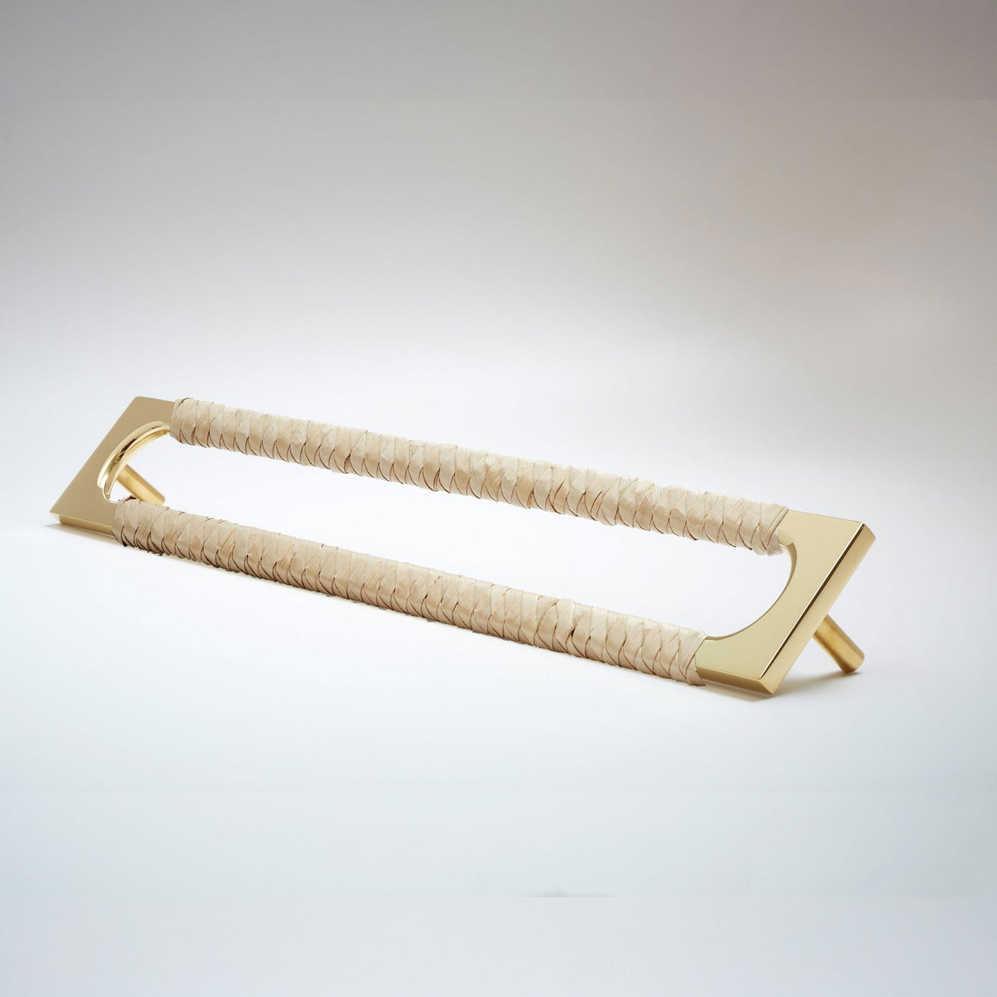 A modern long rectangular cabinet pull handle with a polished brass metal frame with an oval shaped-cutout and a light tan leather woven grip, shown on a white background.