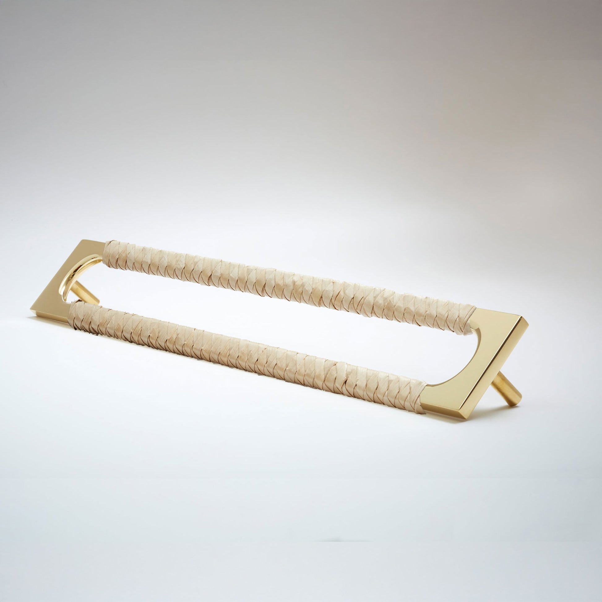 A modern long rectangular cabinet pull handle with a polished brass metal frame with an oval shaped-cutout and a light tan leather woven grip, shown on a white background.