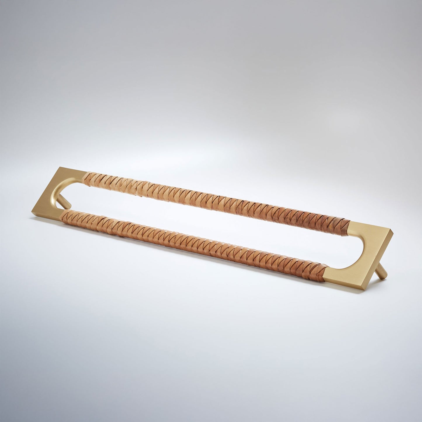 A modern long rectangular cabinet pull handle with a satin brass metal frame with an oval shaped-cutout and a brown leather woven grip, shown on a white background.