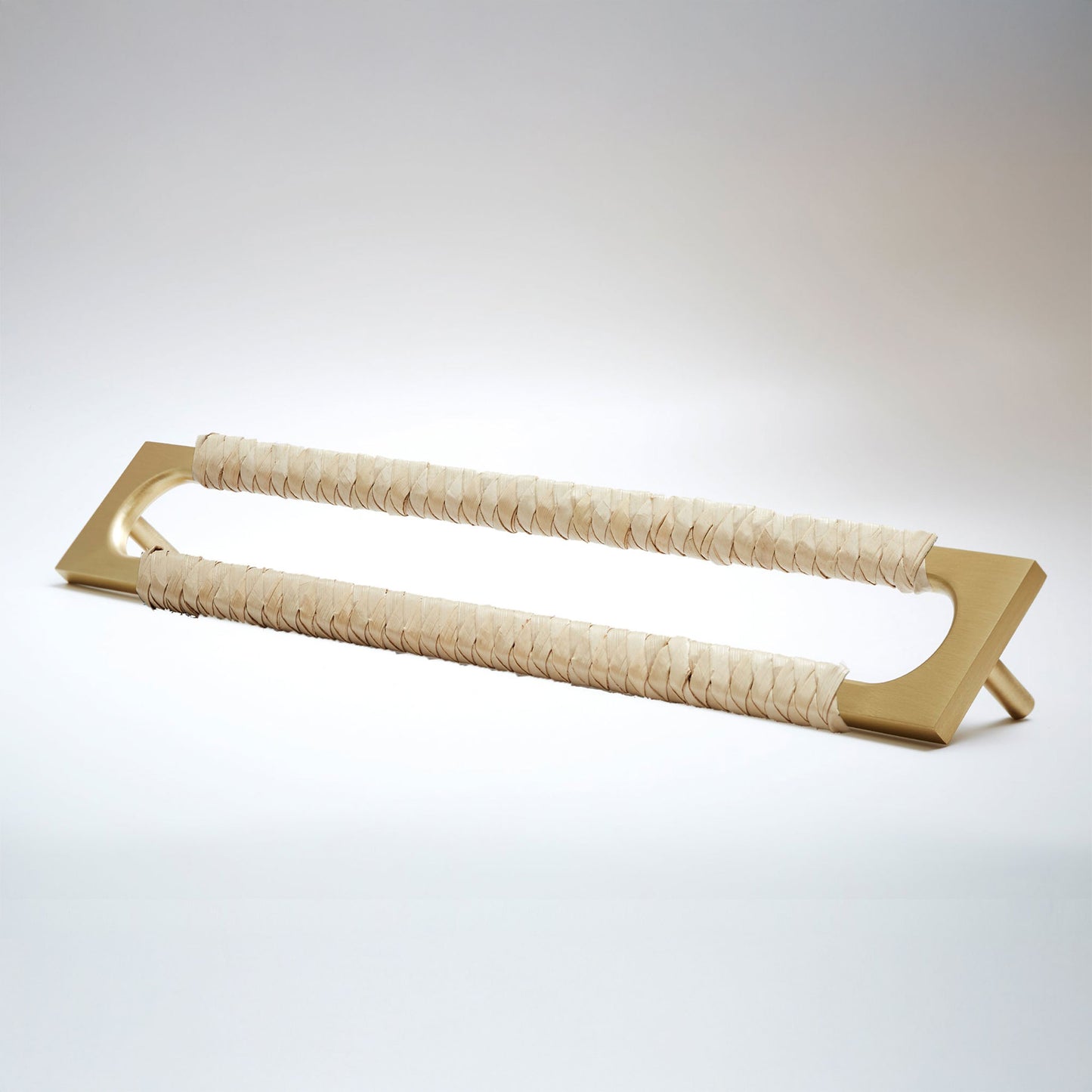 A modern long rectangular cabinet pull handle with a satin brass metal frame with an oval shaped-cutout and a light tan leather woven grip, shown on a white background.