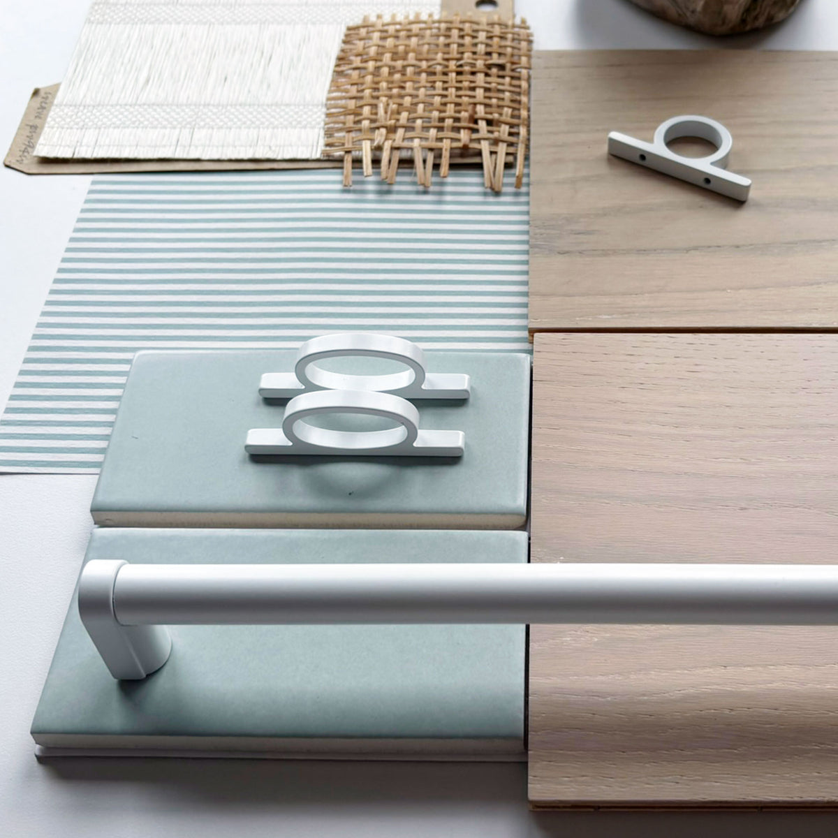 a design flat lay with various textured fabrics and wood finishes and including matte white loop shaped knobs and appliance pulls along side pale sage colored ceramic tiles on a white background.