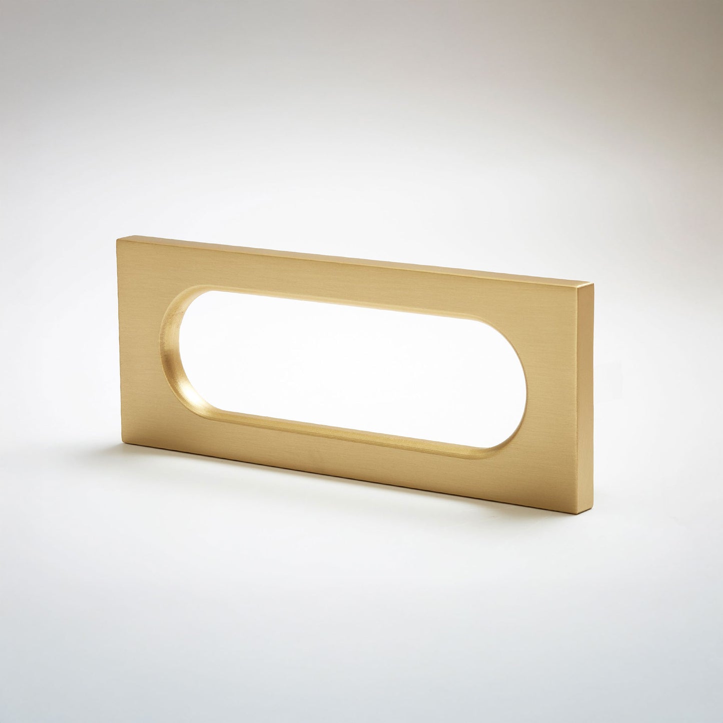 A modern rectangular pull handle with a brushed brass finish and an oval cutout, shown on a clean light gray background.