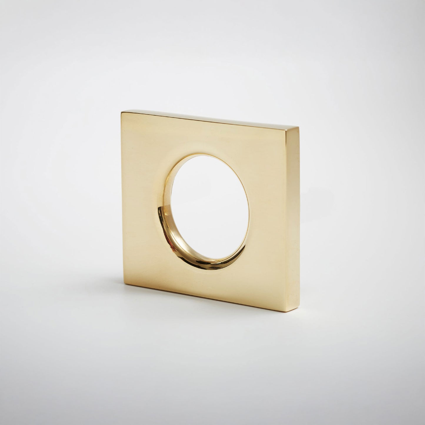 A modern square gold-toned metal cabinet pull with a circular cutout in the center, shown on a clean light gray background.