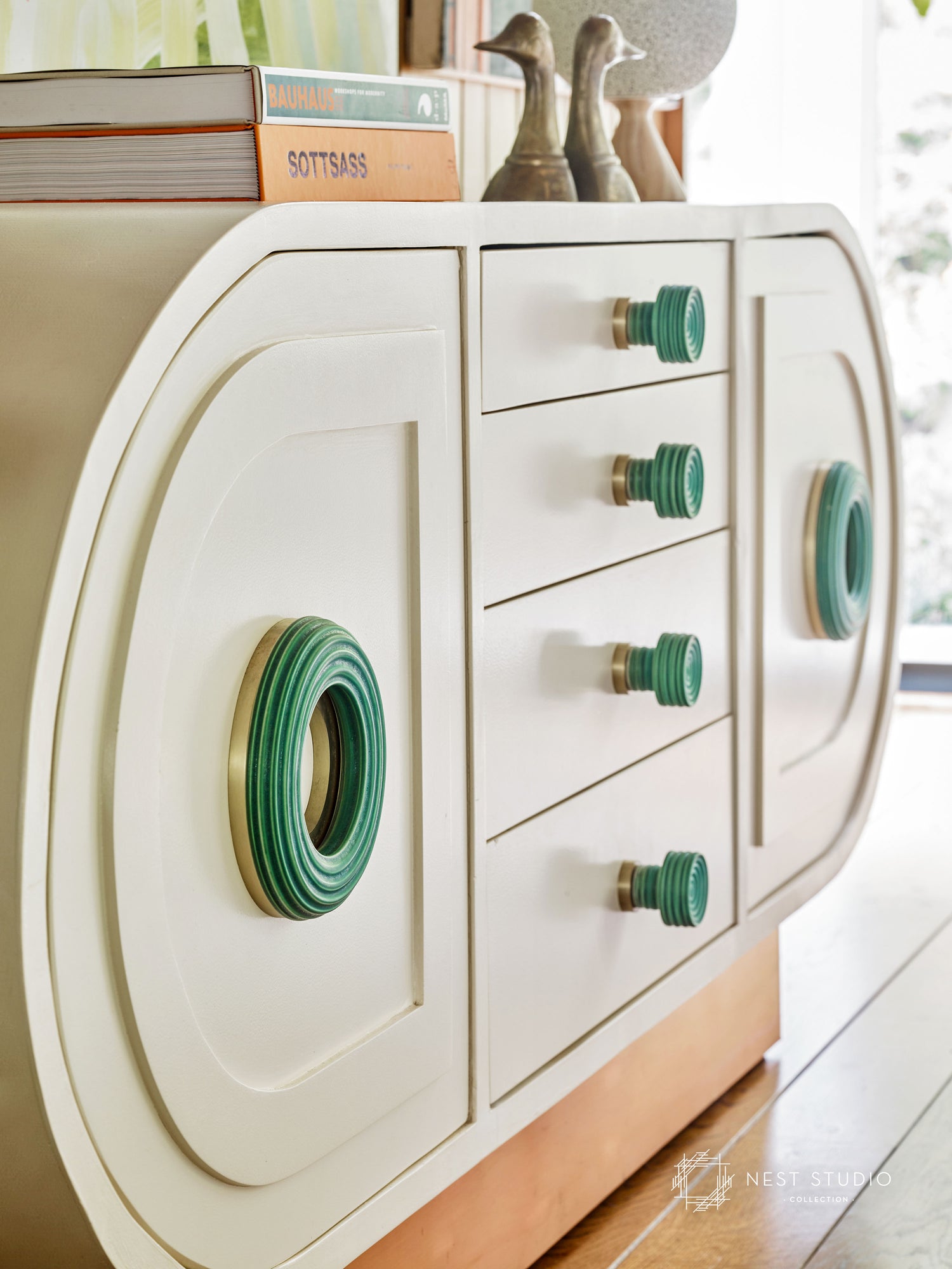 A cream-colored mid-century modern dresser with teal green and gold drawer knobs and matching green ring pulls, shown in a bright room setting.
