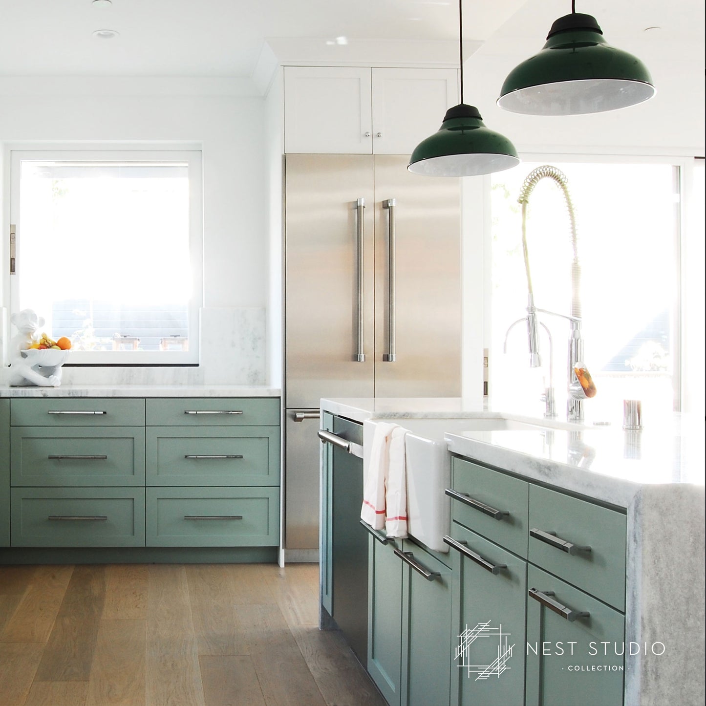 A modern kitchen featuring sage green cabinetry, white marble countertops, a stainless steel refrigerator, and two dark green pendant lights. A white towel with red stripes hangs on the cabinet handle, and a 'NEST STUDIO COLLECTION' watermark is visible in the bottom right corner.