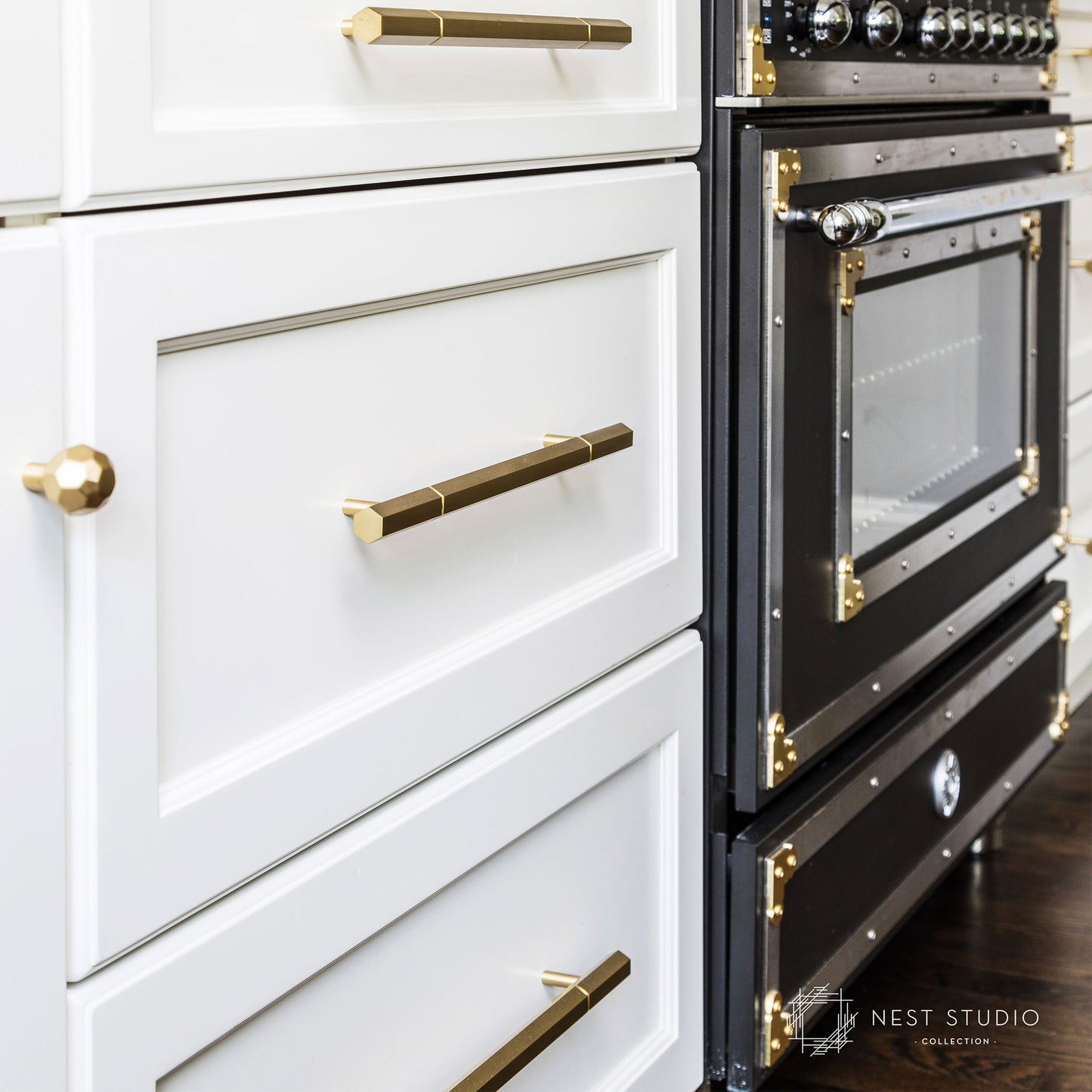 Close-up of a modern kitchen cabinet and built-in oven. The white cabinets feature brass and gold hardware, including a round knob on the left drawer and long bar handles on the drawers and oven door.