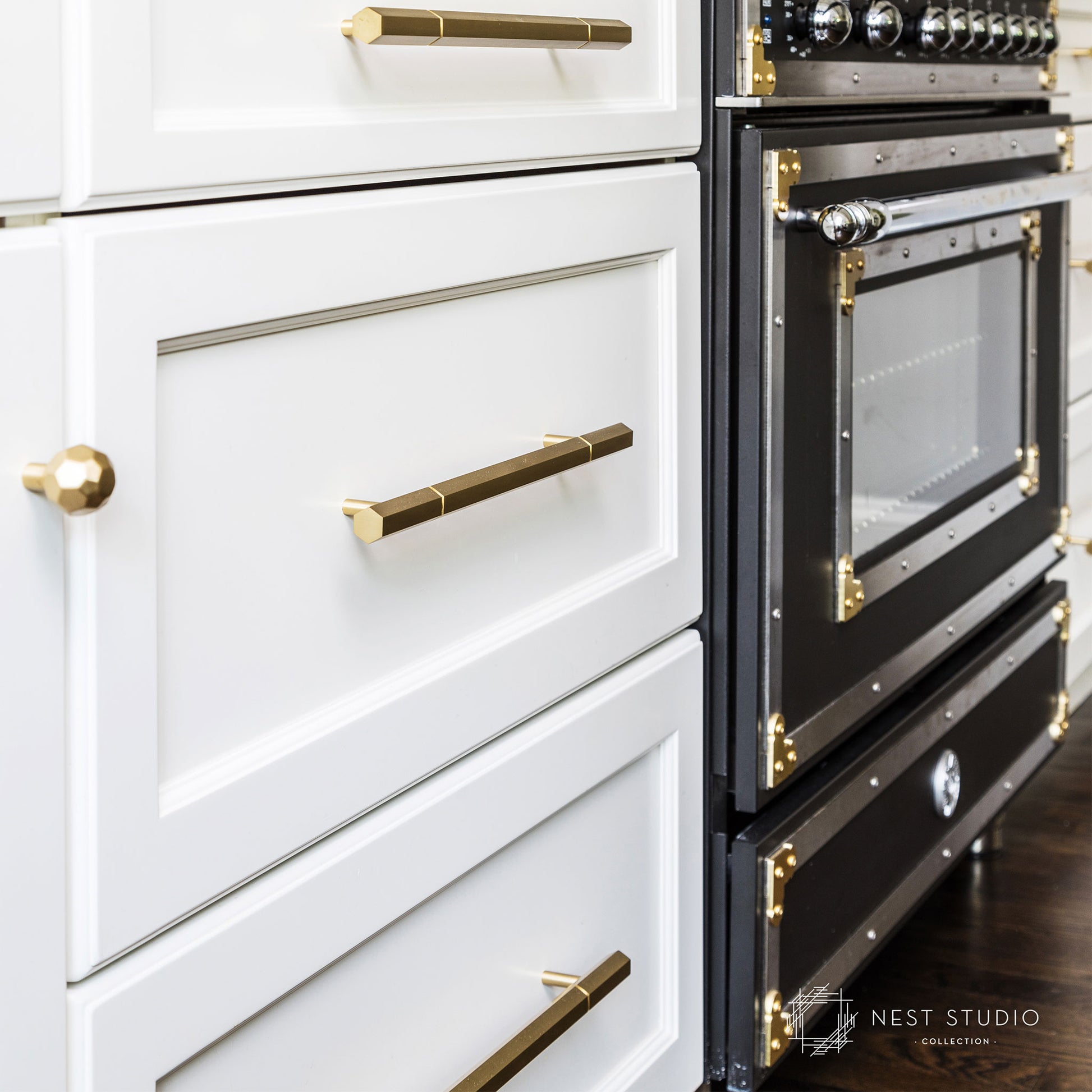 Close-up of a modern kitchen cabinet and built-in oven. The white cabinets feature brass and gold hardware, including a round knob on the left drawer and long bar handles on the drawers and oven door.