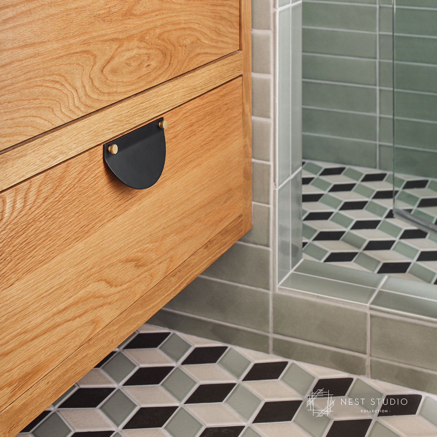 Close-up of a wooden vanity cabinet with a black metal pull handle in a modern bathroom, next to a glass shower door and patterned tile floor.