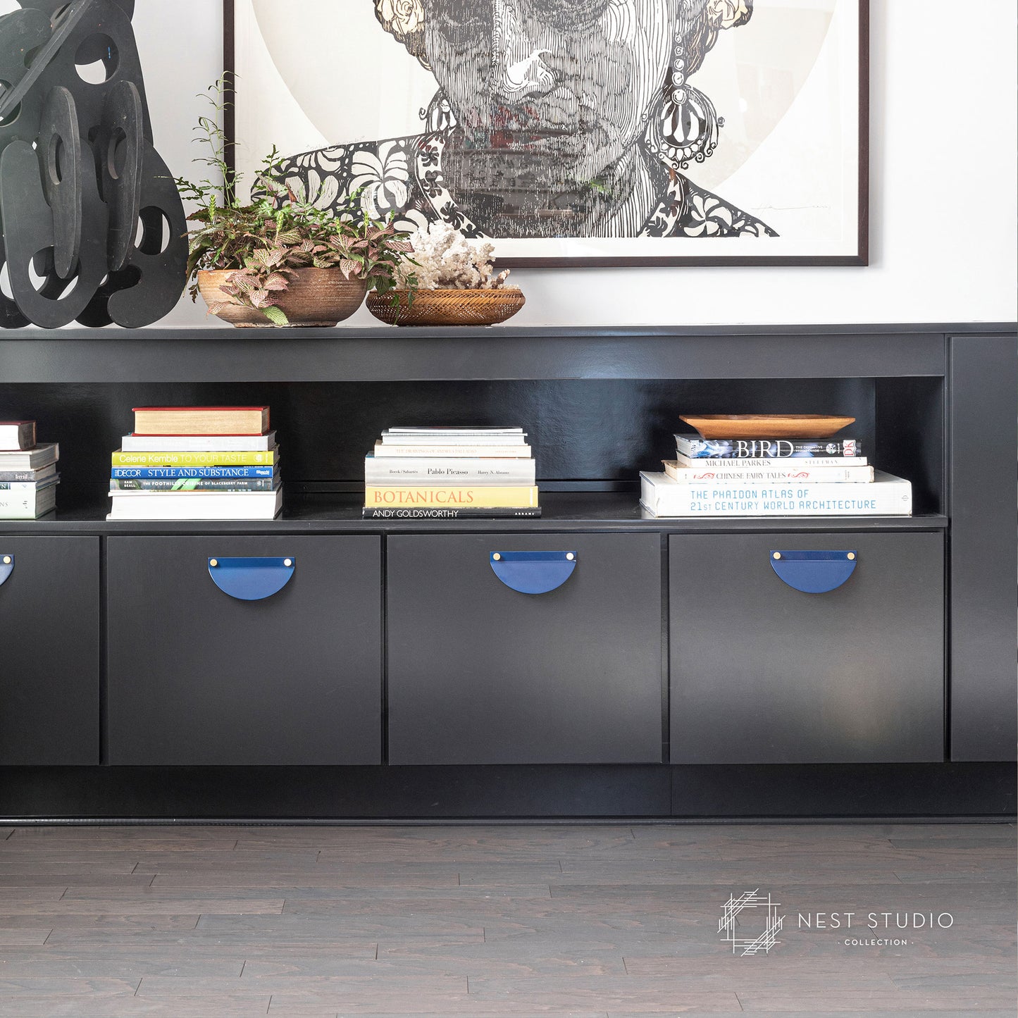 A modern black media console with open shelving and four cabinet doors featuring blue half oval pulls, styled with books, potted plants, and a framed artwork above, set against a white wall and dark wood flooring.