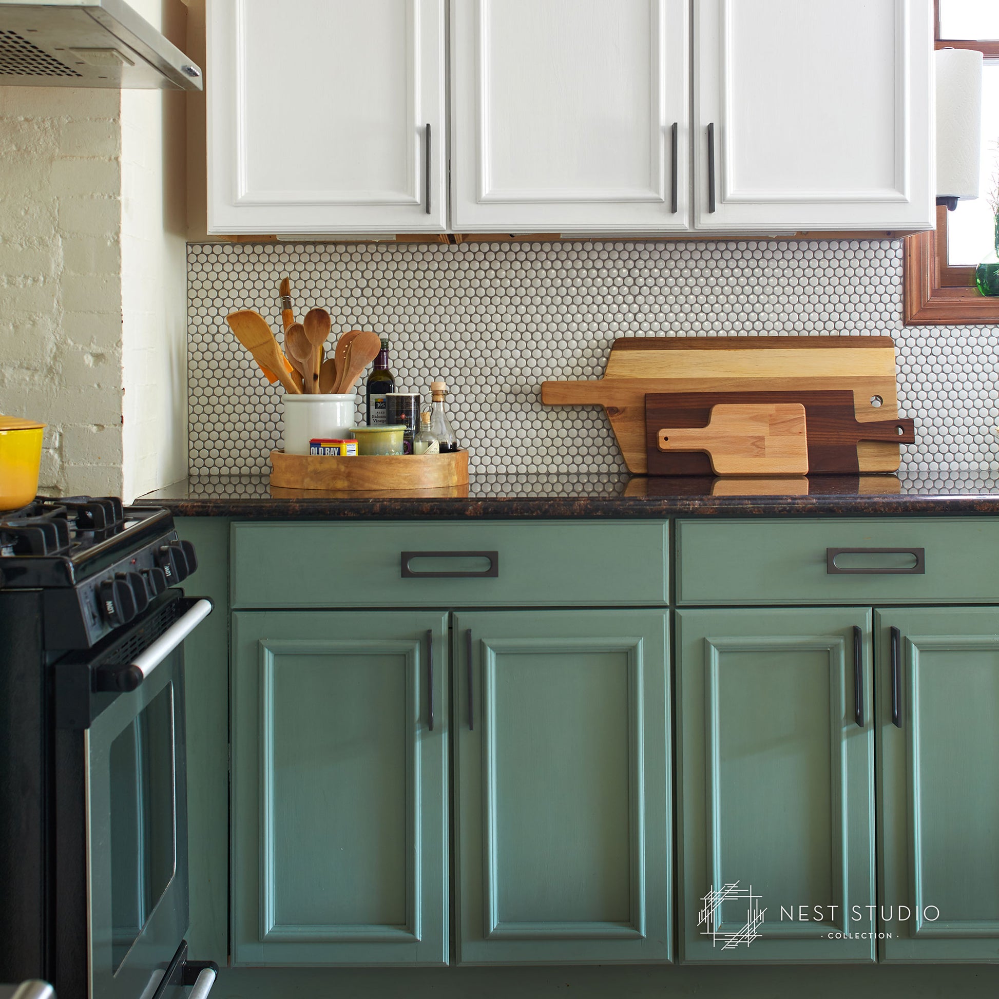 A kitchen scene with sage green lower cabinets, white upper cabinets, a small black-and-white hexagon tile backsplash, and wooden cutting boards on the countertop. A black stove is partially visible on the left, and a Nest Studio Design watermark appears in the lower right corner.