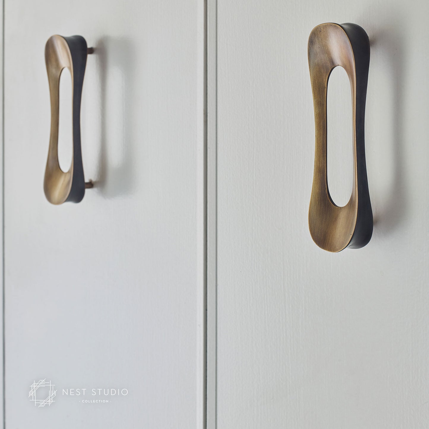 Two modern elongated cabinet pulls in a brushed brass or bronze finish with dark backing, mounted on white cabinet doors. A Nest Studio watermark is visible in the lower-left corner.