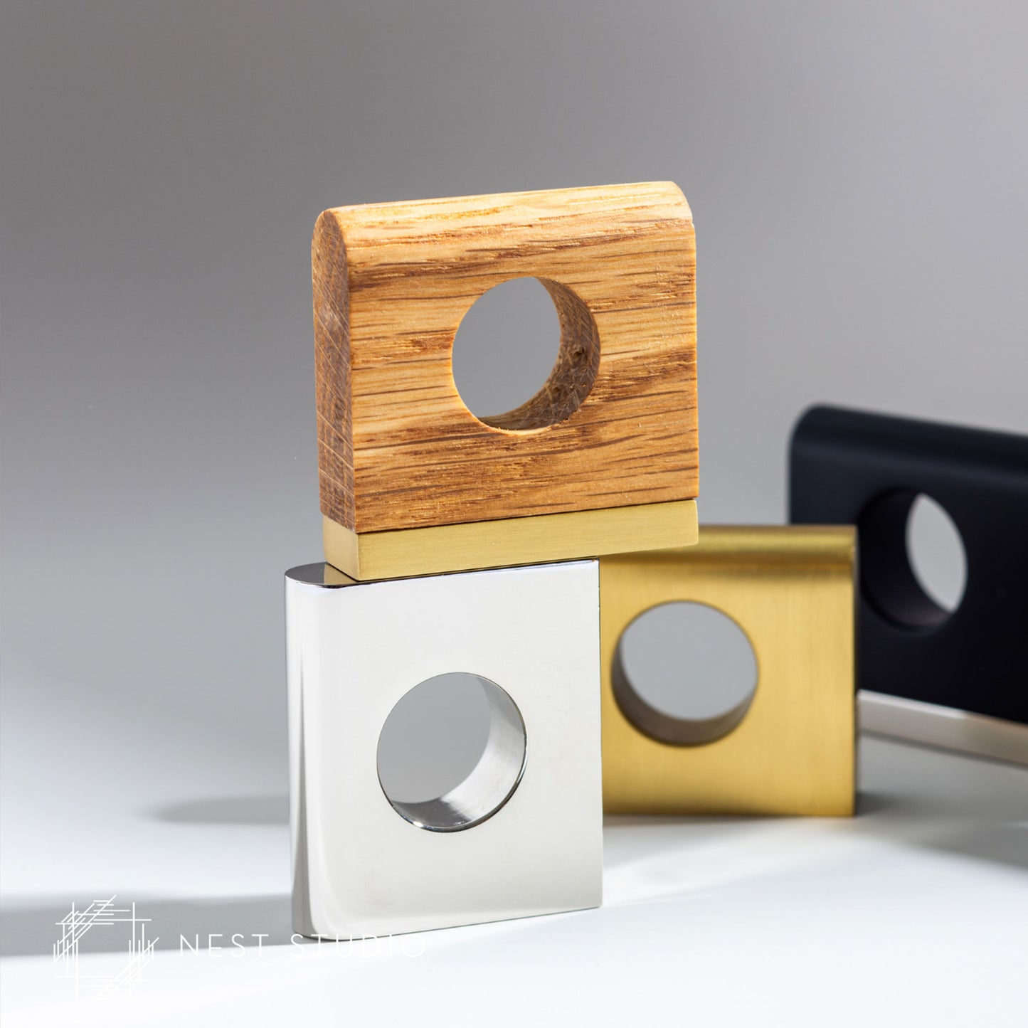 Three modern square cabinet pulls with circular cutouts are stacked on a light gray surface against a neutral gray background. The top holder has a natural wood grain top and a brushed gold base, the middle holder has a brushed silver top and a gold base, and a third black holder is partially visible in the background.
