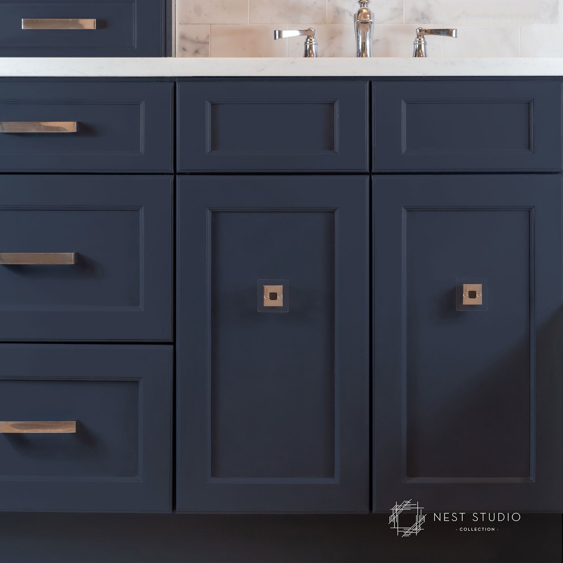 A close-up of a modern bathroom vanity cabinet in a deep navy blue finish, featuring recessed panel doors and drawers with brushed brass hardware and a white countertop.