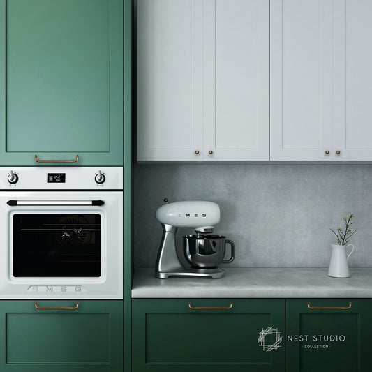 A modern kitchen scene featuring sage green and white cabinets, a built-in oven, a silver stand mixer on a gray countertop, and a small white vase with dried stems. The cabinets have brass handles, and a 'Nest Studio Collection' logo is visible in the bottom right corner.