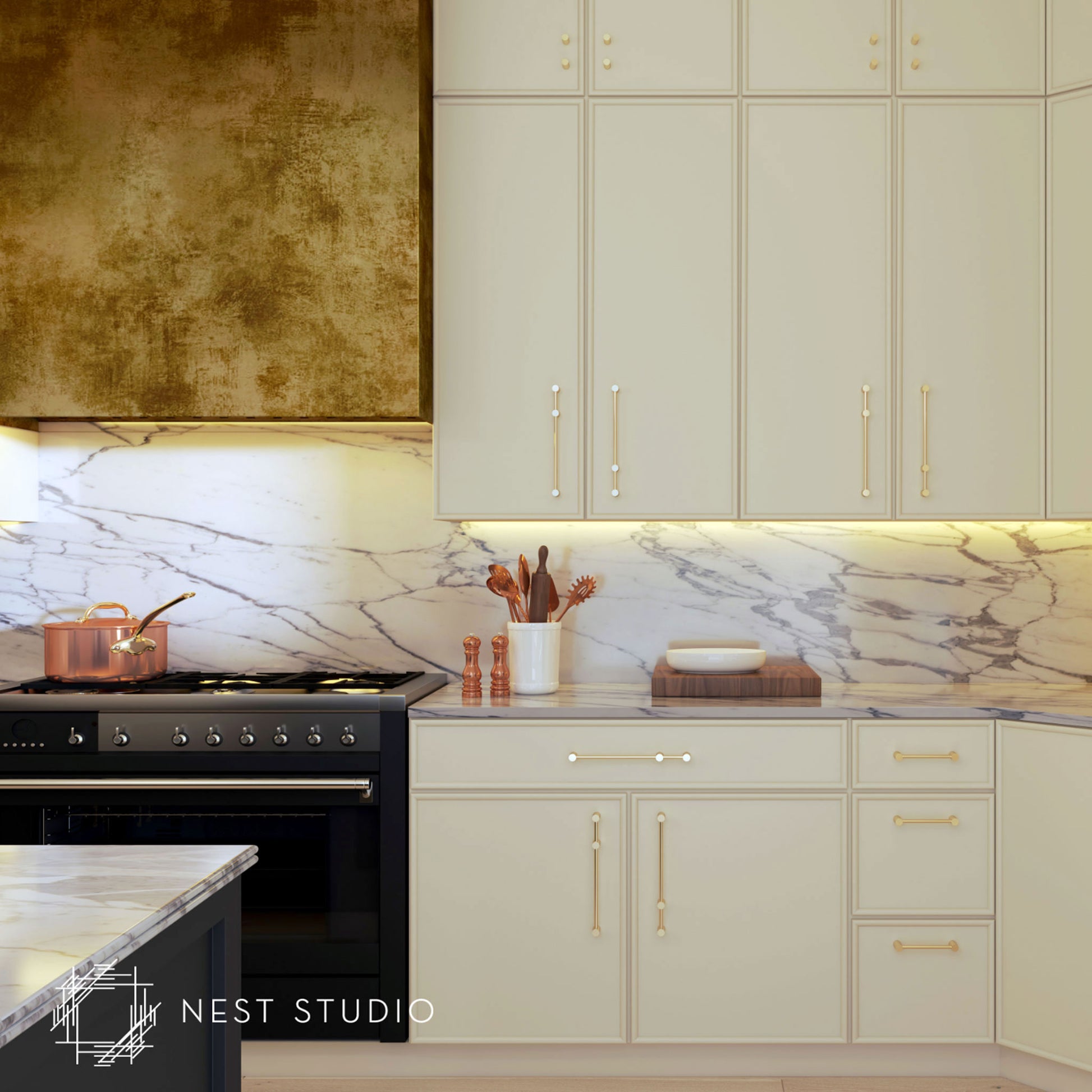 Cream-colored kitchen cabinets with gold handles and a white marble backsplash, featuring a black stove and a textured gold range hood. The Nest Studio logo is visible in the bottom-left corner.