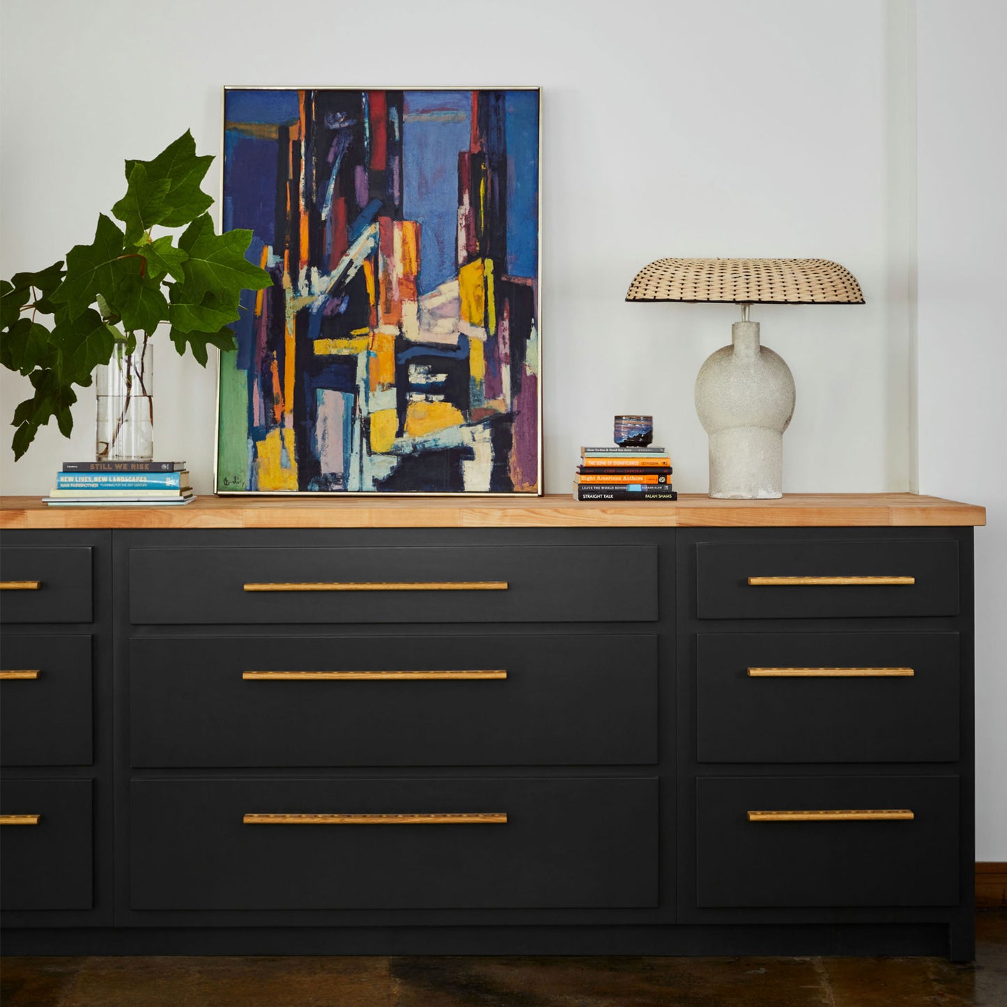 A modern black wooden sideboard or dresser with gold-toned metal handles, styled in a room setting with a light wood top, a potted plant, books, a lamp, and a colorful abstract painting.