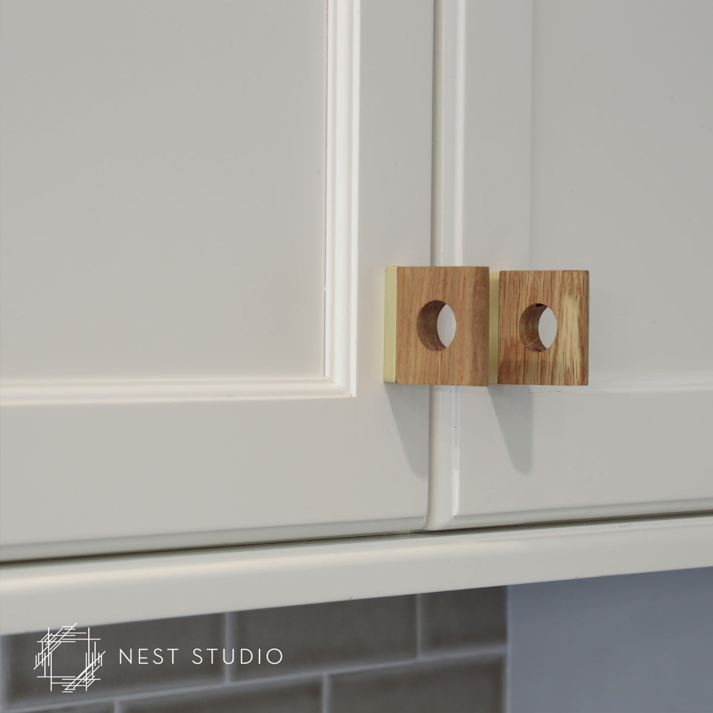 Two square wooden cabinet pulls with circular cutouts, mounted on white kitchen cabinets. A NEST STUDIO watermark is visible in the bottom-left corner.