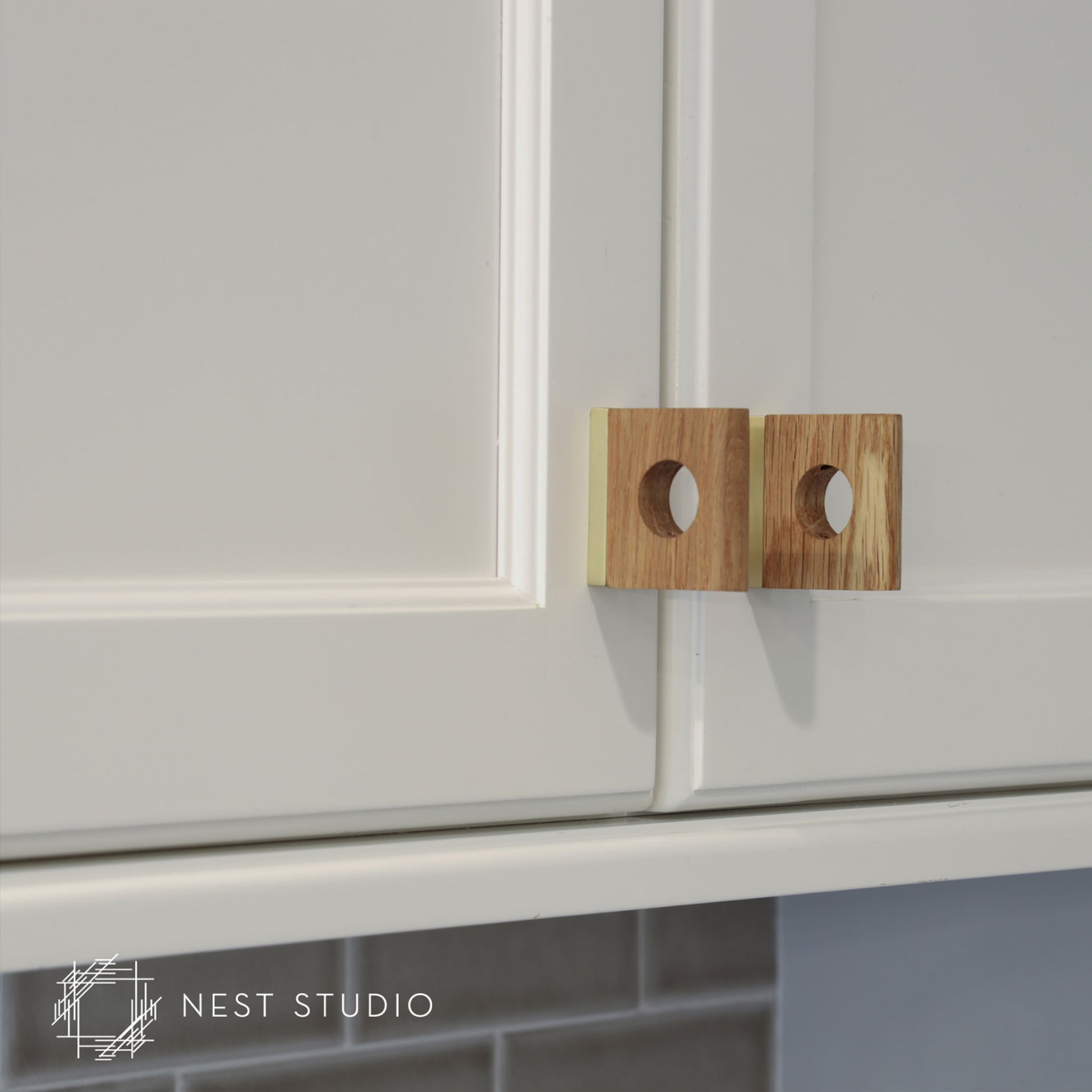 Two square wooden cabinet pulls with circular cutouts, mounted on white kitchen cabinets. A NEST STUDIO watermark is visible in the bottom-left corner.