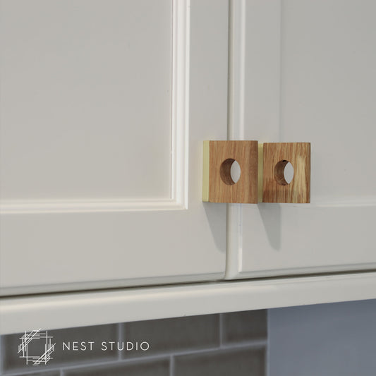 Two square wooden cabinet pulls with circular cutouts, mounted on white kitchen cabinets. A NEST STUDIO watermark is visible in the bottom-left corner.