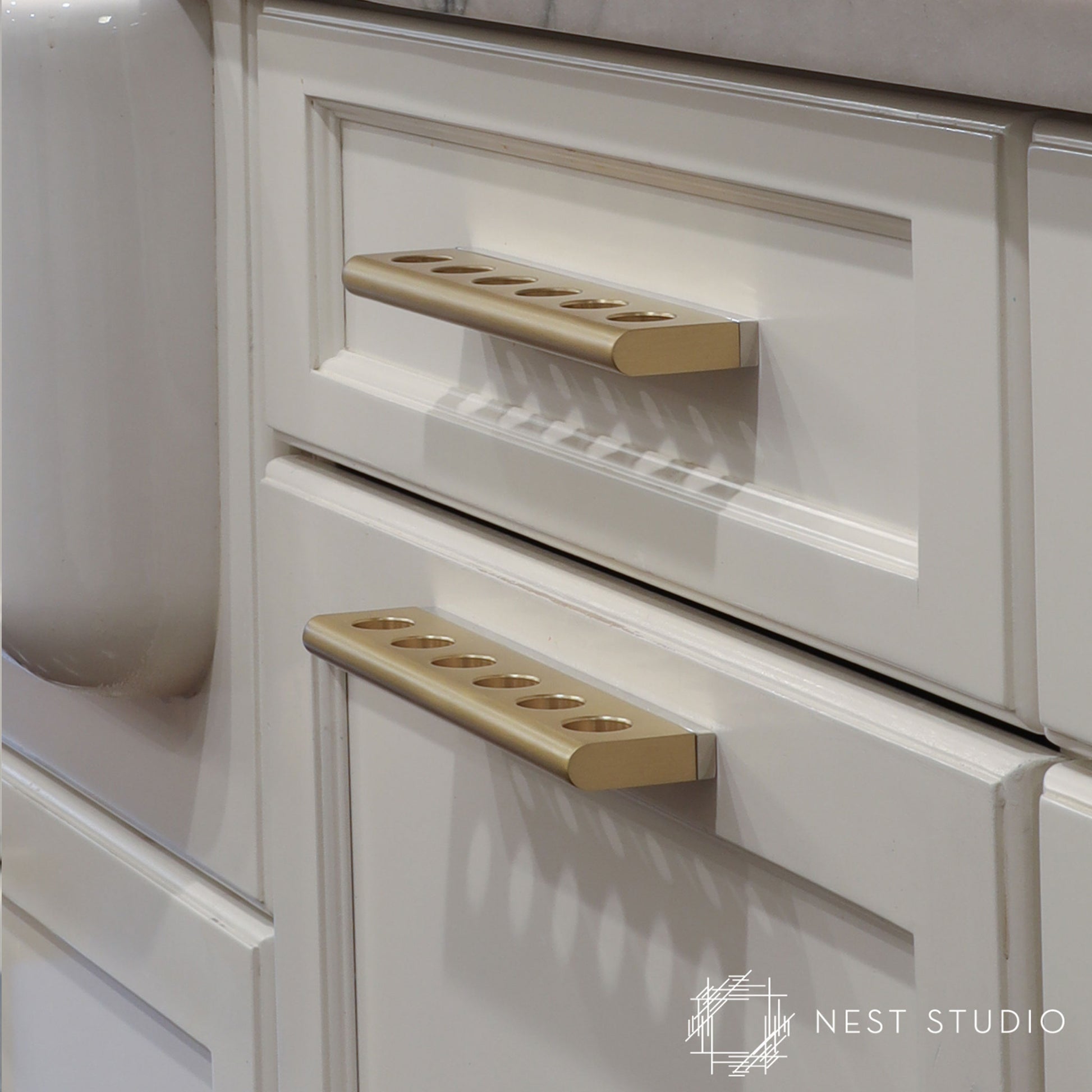 Close-up of a cream-colored kitchen cabinet with two drawers, each fitted with a modern rectangular pull handle in a warm brushed gold finish. The cabinet features raised panel detailing and a light stone countertop is partially visible at the top.