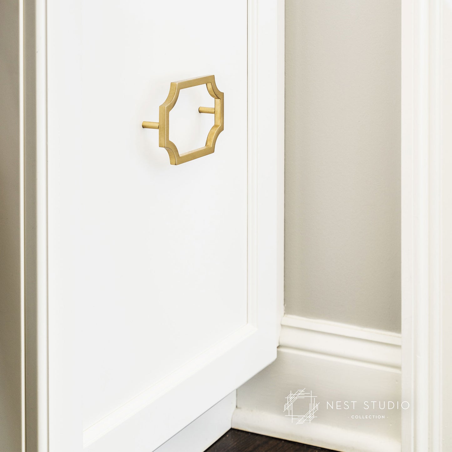 A close-up of a white cabinet door featuring a decorative gold octagonal pull handle, with a glimpse of a beige wall and baseboard on the right. A 'Nest Studio Collection' watermark is visible in the bottom right corner.