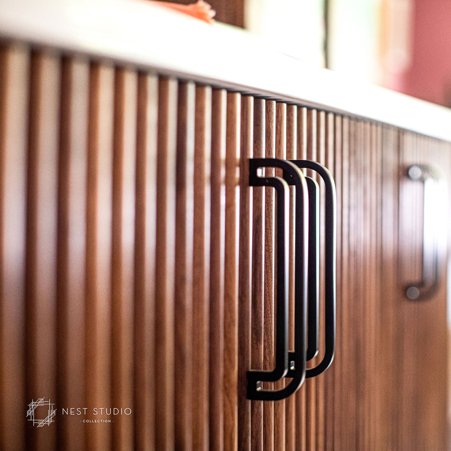 A close-up of a modern kitchen cabinet featuring vertical wood slats and a bold black metal handle, with a second matching handle visible in the background.