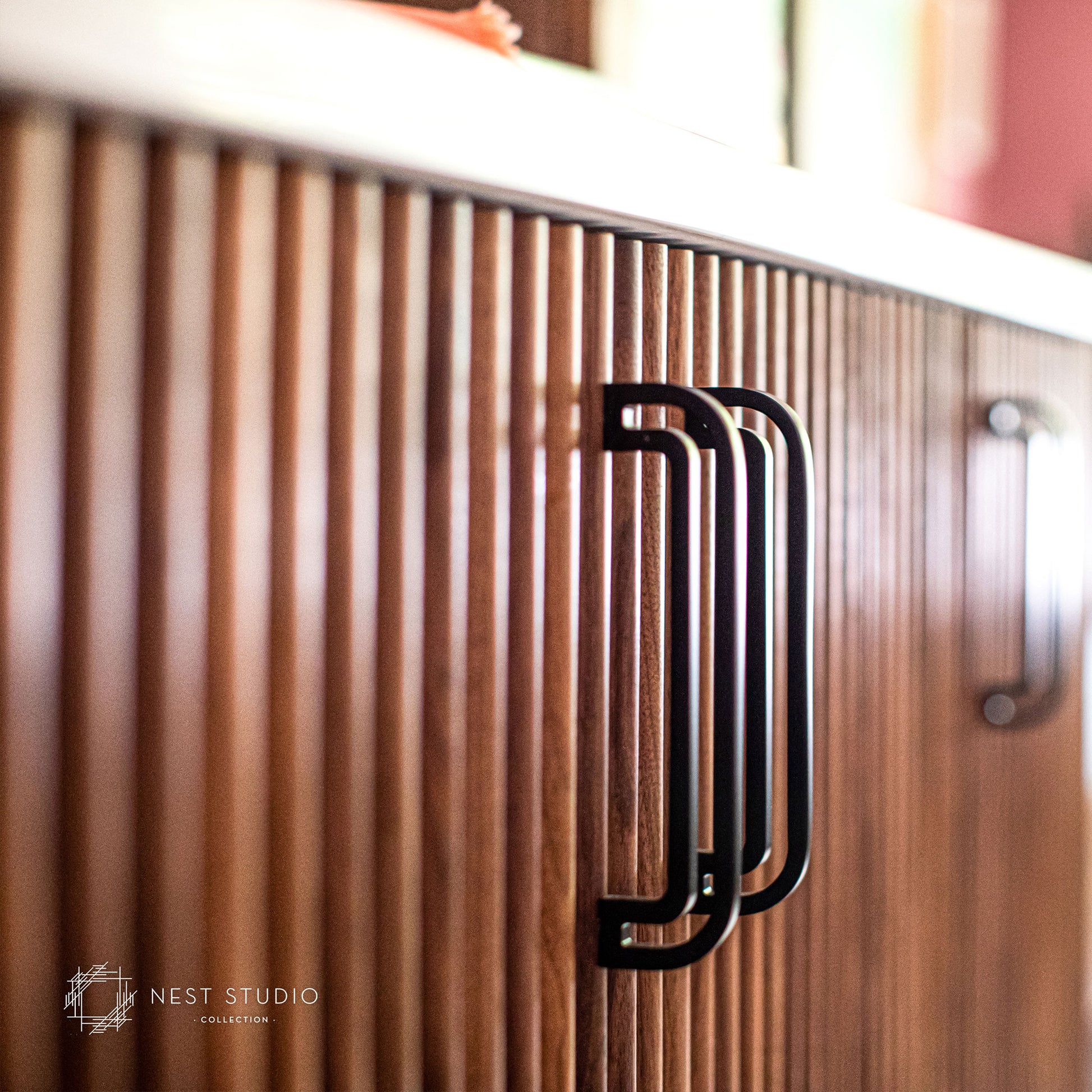 A close-up of a modern kitchen cabinet featuring vertical wood slats and a bold black metal handle, with a second matching handle visible in the background.