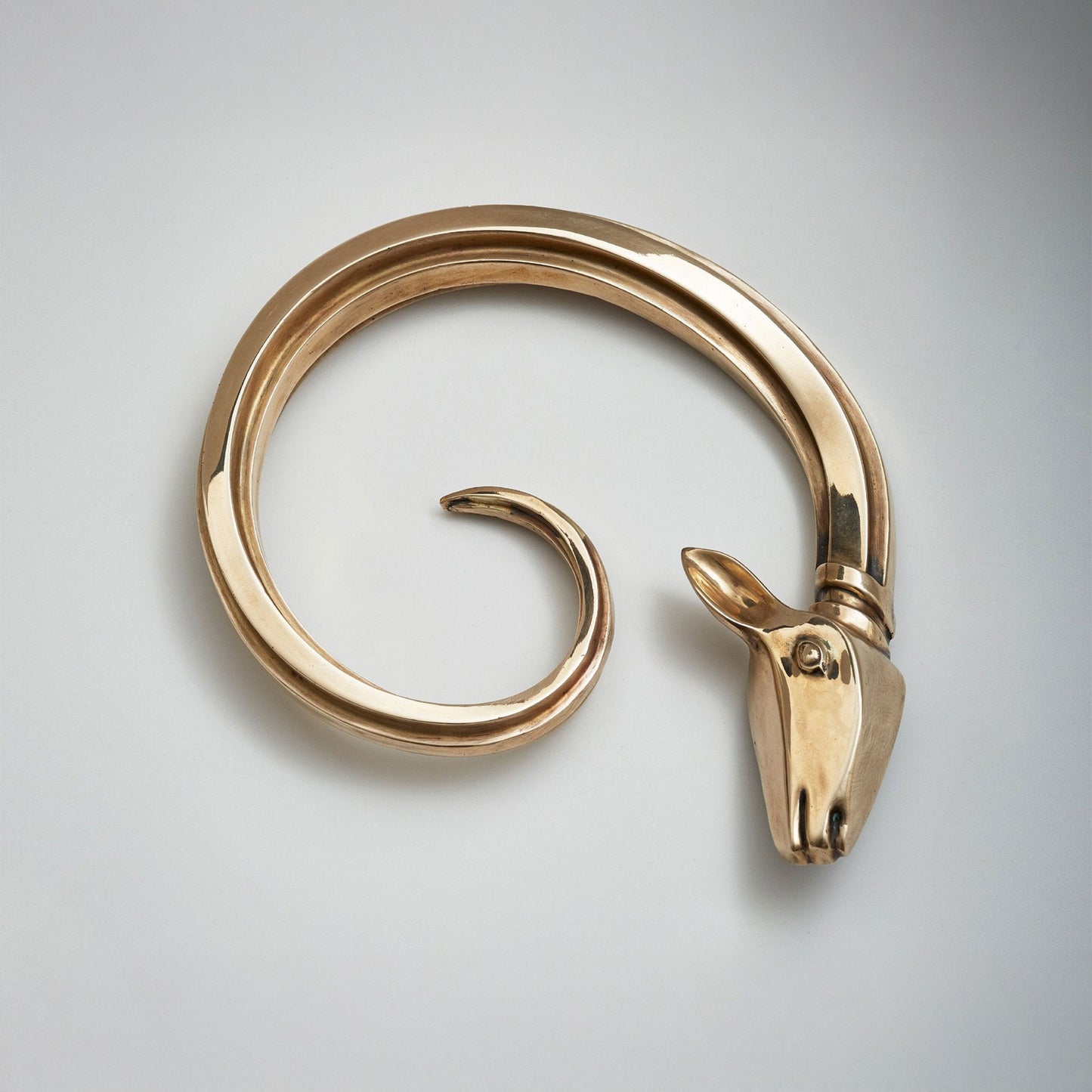 A polished brass-toned metal spiral cabinet hardware pull with a sculpted ram head at one end, on a clean light gray background.