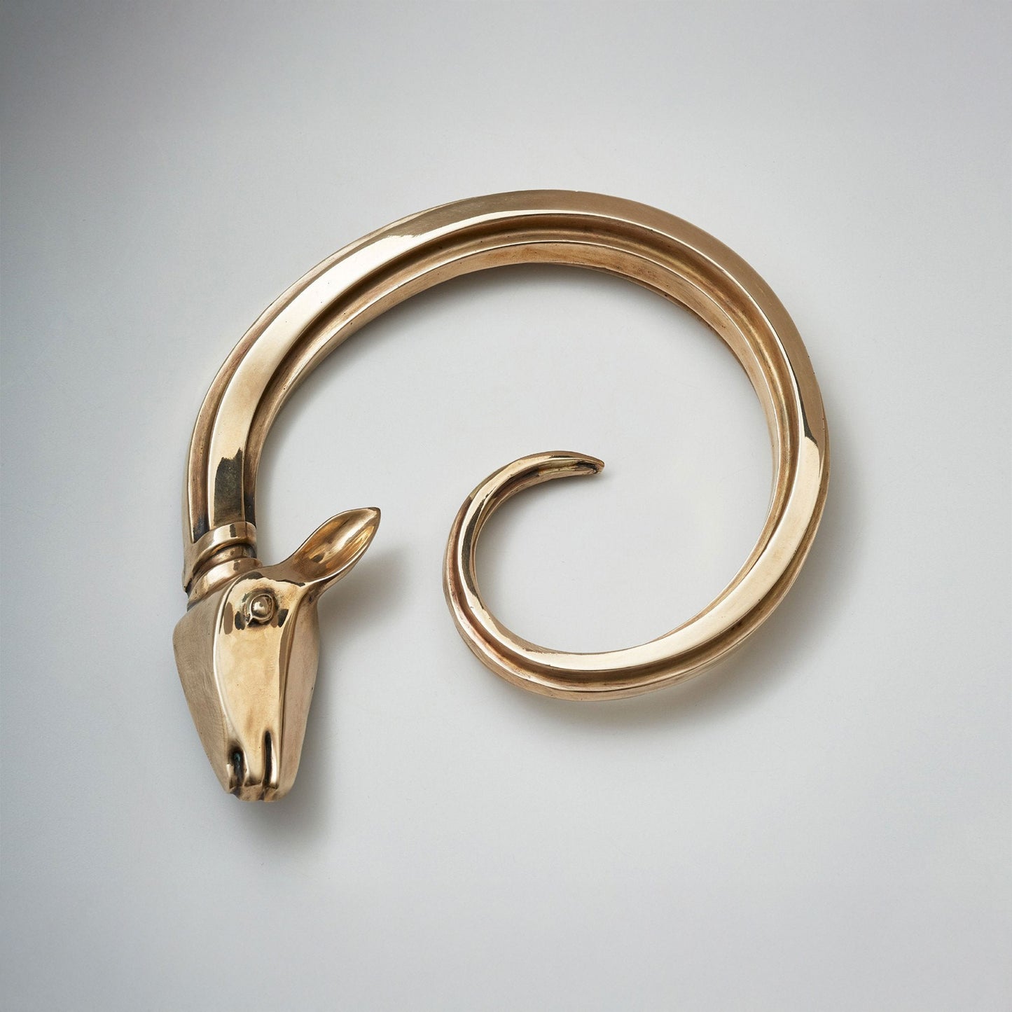 A polished brass-toned metal spiral cabinet hardware pull with a sculpted ram head at one end, on a clean light gray background.