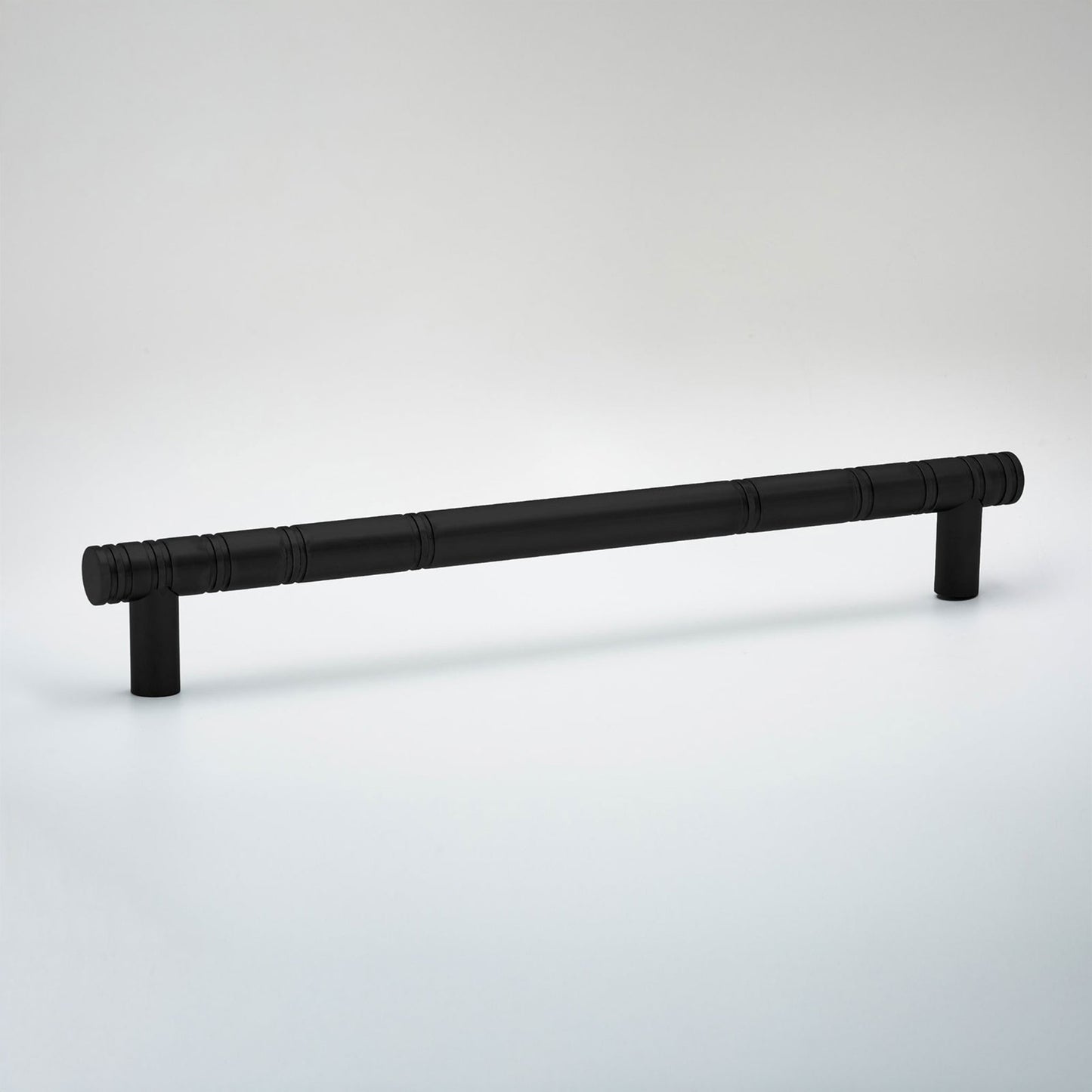 A matte black metal cabinet pull handle with a textured, ribbed cylindrical shaft and two short post bases, shown on a light gray gradient studio background.