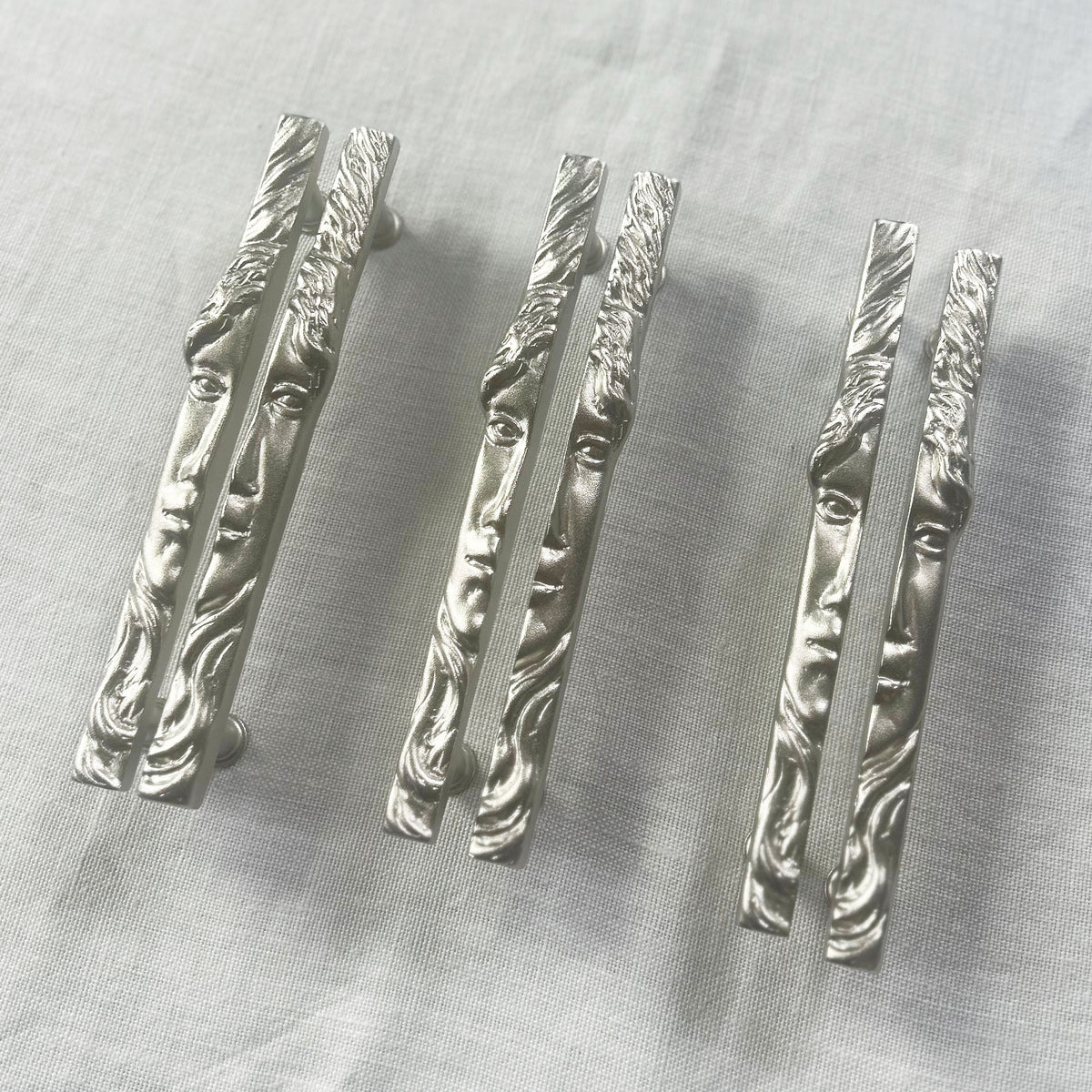 A grouping of three sets of face shaped cabinet pull pairs in matte nickel on a natural woven background