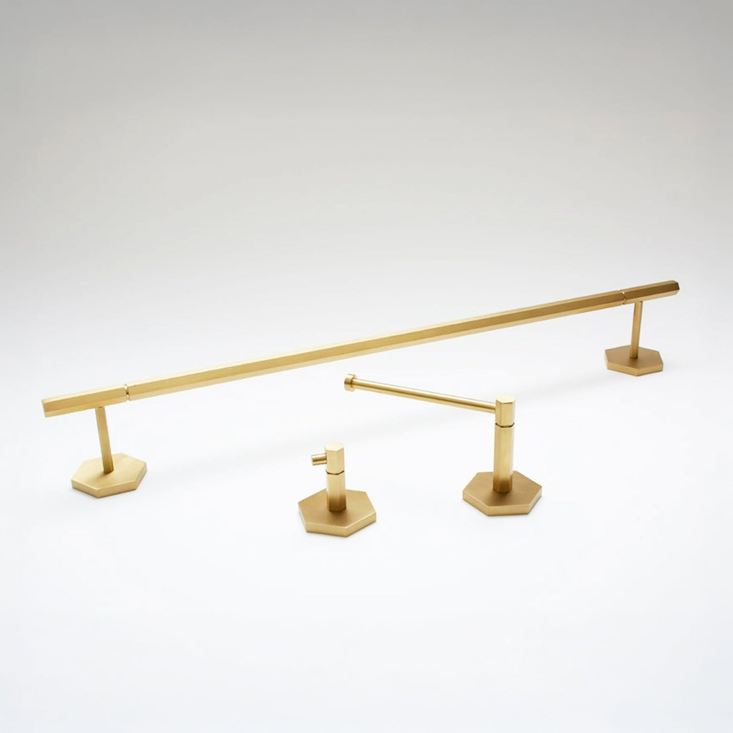 A set of modern brass bathroom fixtures, including a long horizontal bar, a shorter vertical bar, and two hexagonal base plates, shown on a light gray gradient background.