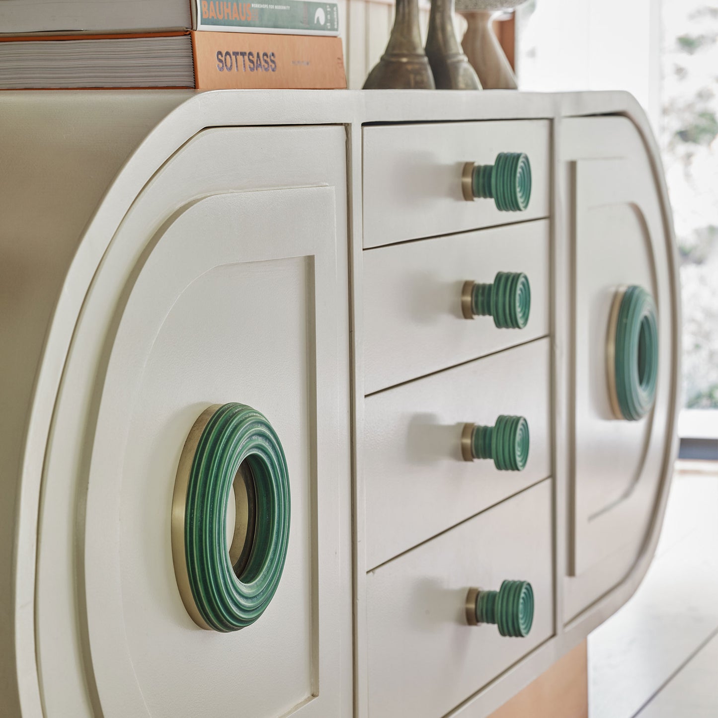 A close-up of a cream-colored dresser with distinctive green and gold knobs, set in a bright room with a window in the background.