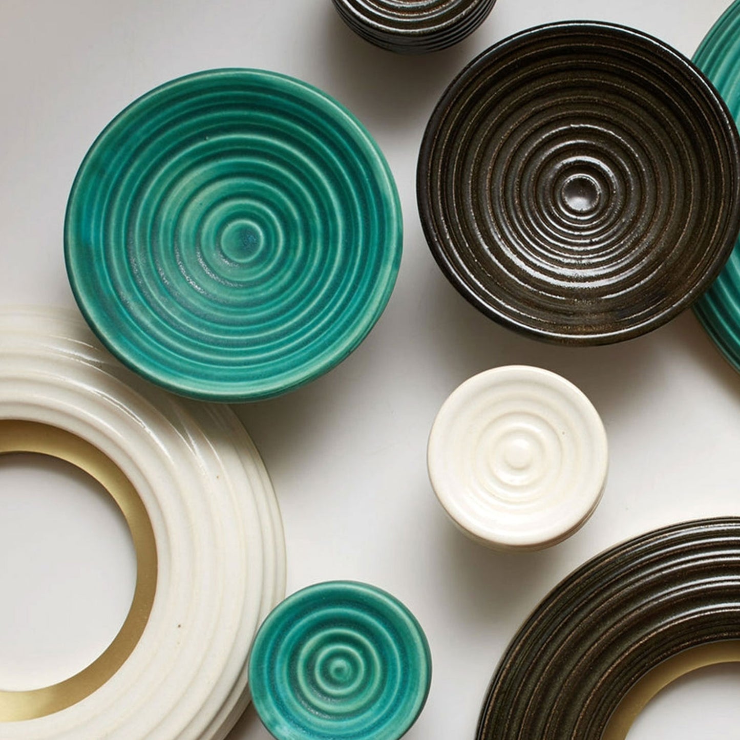 A collection of handmade ceramic cabinet pulls with concentric spiral ridges, shown in teal, dark brown, cream, and white on a light gray background.