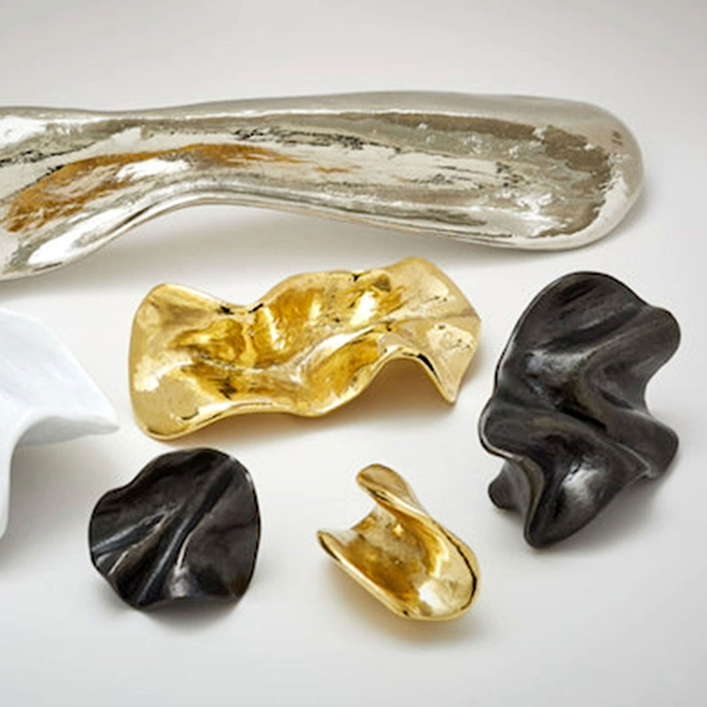 A set of abstract sculptural cabinet pulls with organic, wavy forms in polished silver, gold, and dark black finishes, arranged on a light gray background.