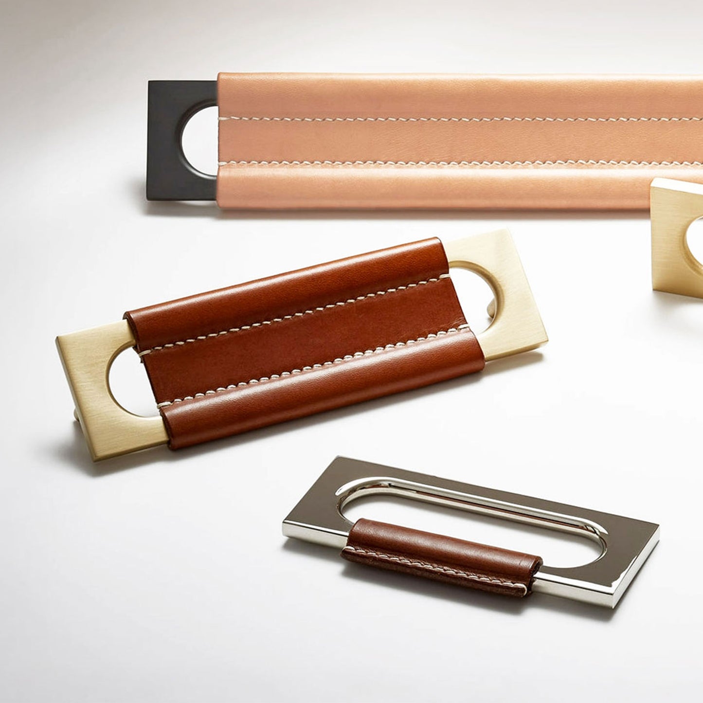 Three leather and metal pull handles arranged on a clean white background. The handles feature stitched leather straps in tan, brown, and dark brown, paired with polished brass and silver-toned metal end pieces.