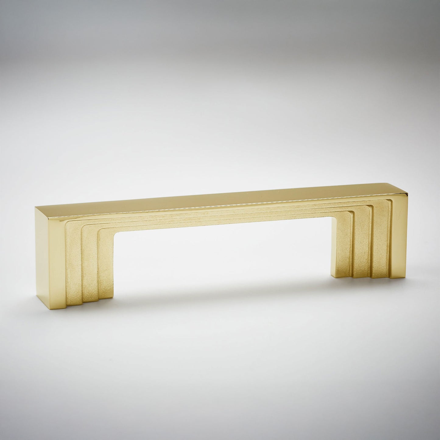 A modern rectangular cabinet pull handle with a brushed gold finish, featuring a stepped geometric design, shown on a neutral light gray background.