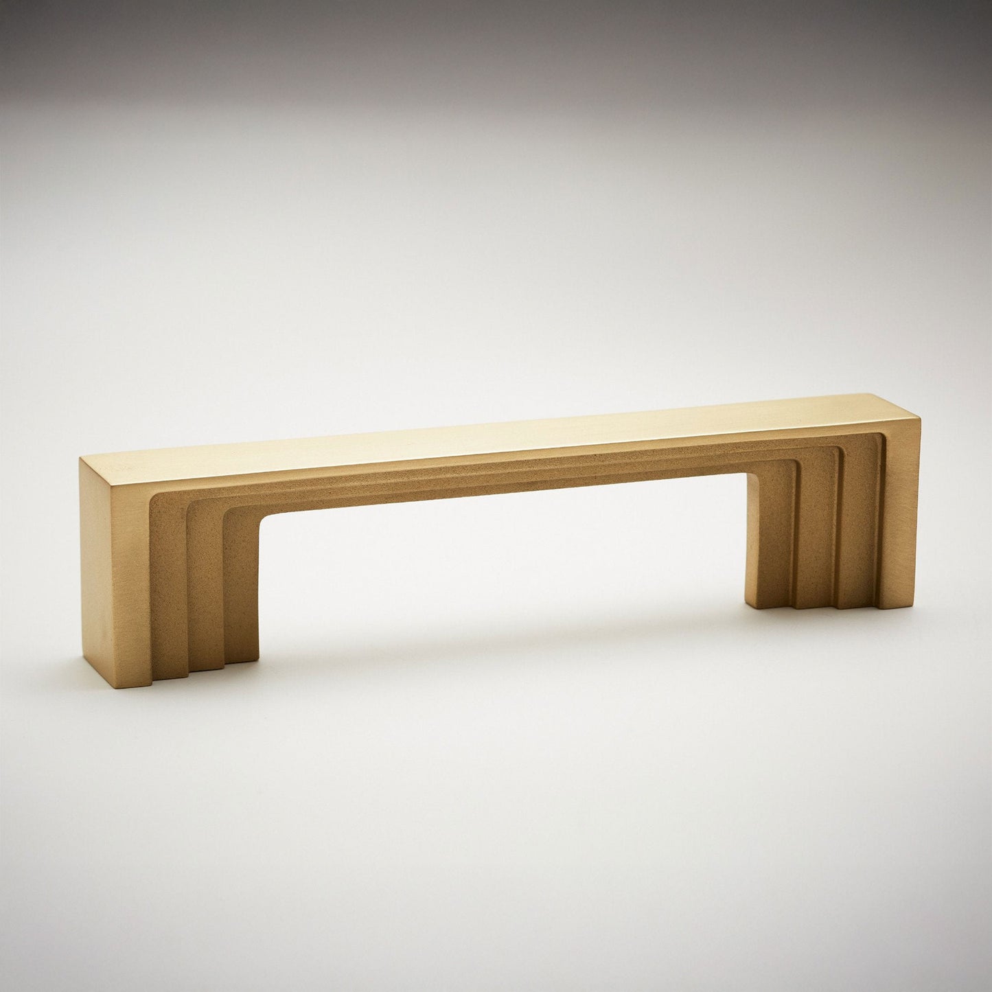 A modern rectangular cabinet pull handle with a warm brushed gold finish, featuring a stepped geometric design and a flat top surface, shown on a neutral light gray background.