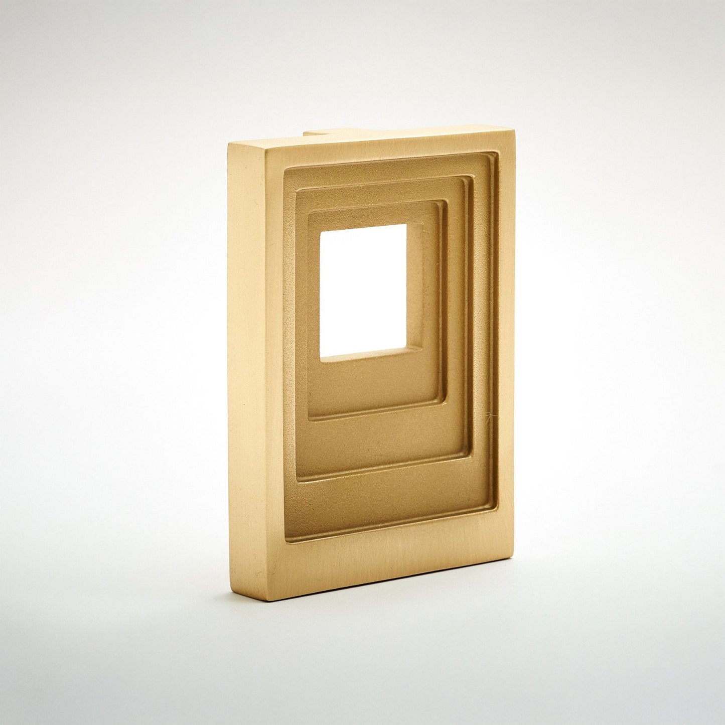 A modern rectangular cabinet pull with a geometric, layered frame design in brushed gold metal, casting a soft light into the center void, set against a clean white background.