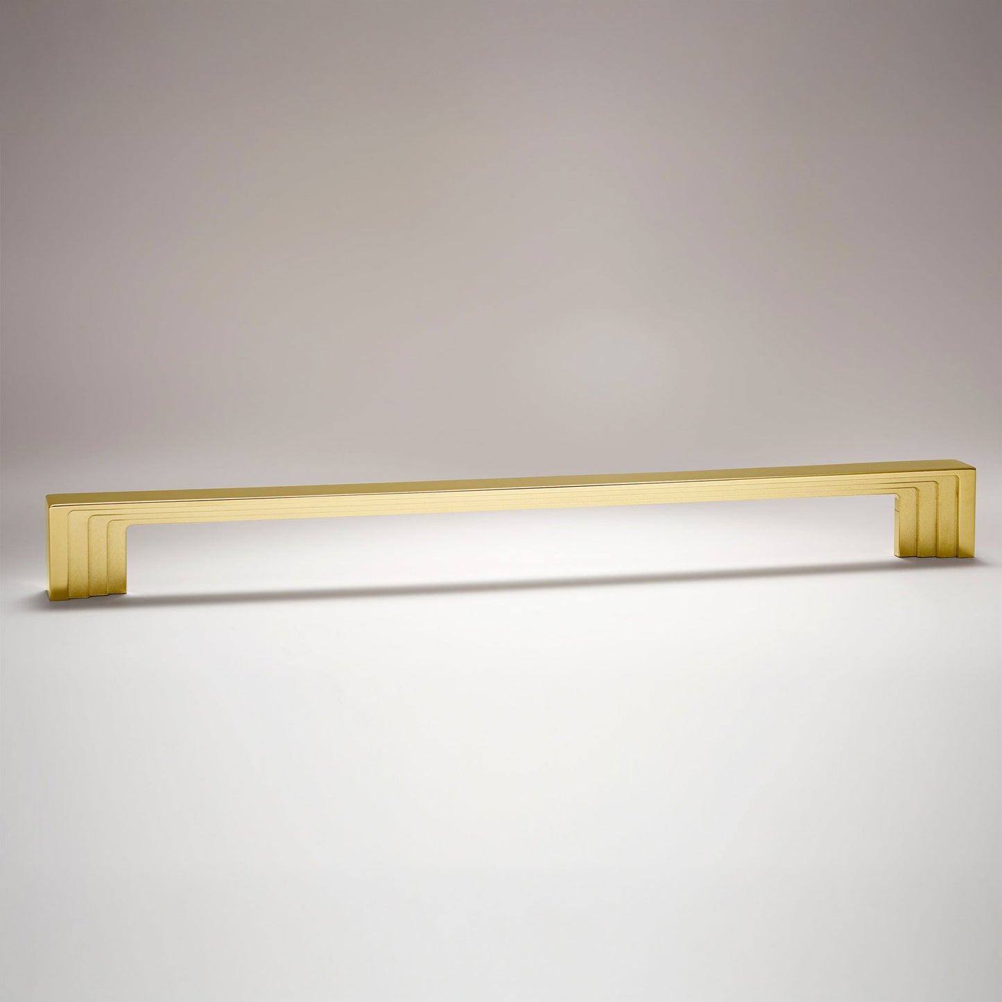 A long, rectangular cabinet pull handle with a brushed gold finish, featuring a modern stepped design and subtle beveled edges, shown against a neutral light gray background.