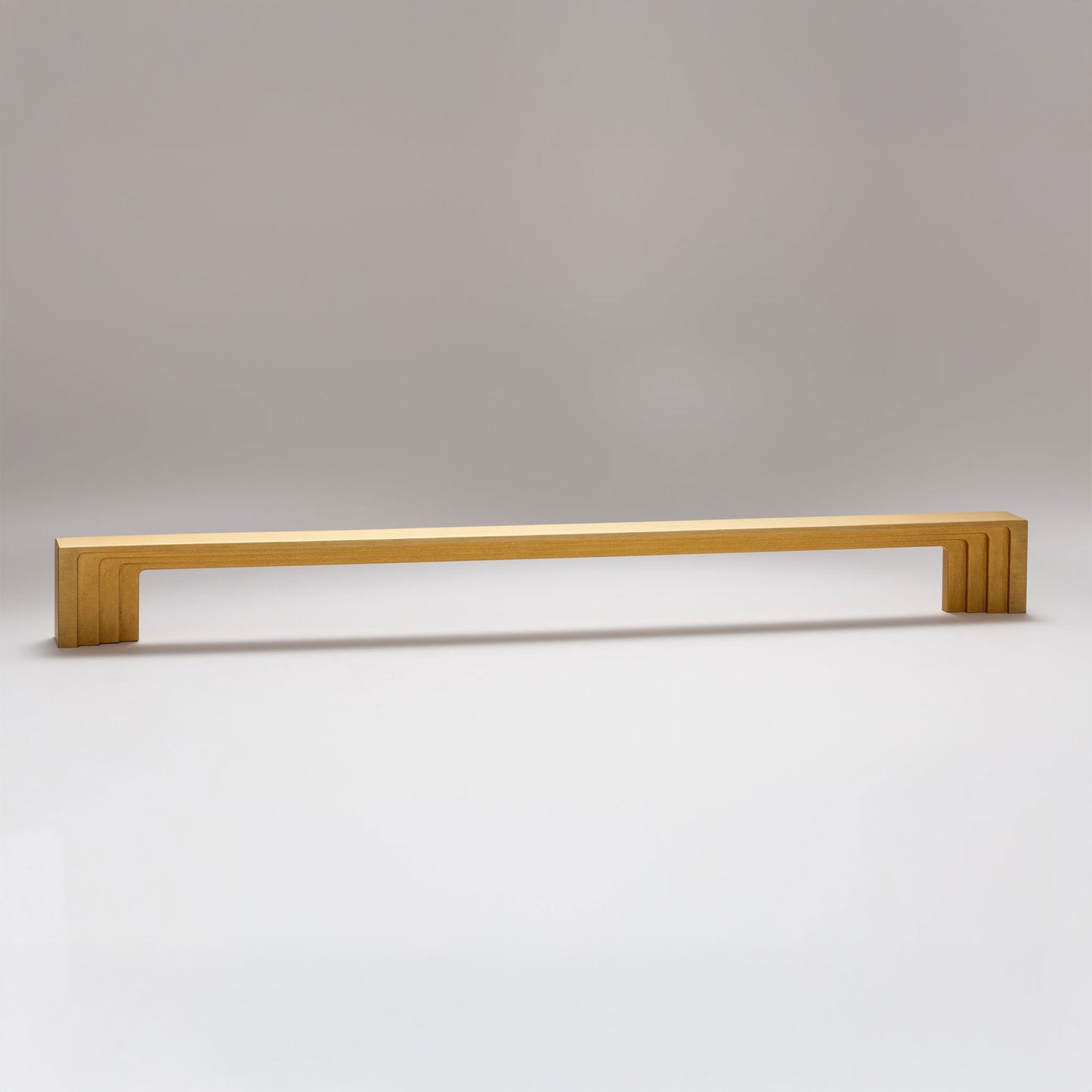 A long, rectangular cabinet pull handle with a brushed gold finish, featuring a modern stepped design and fluted ends, shown against a neutral gray background.