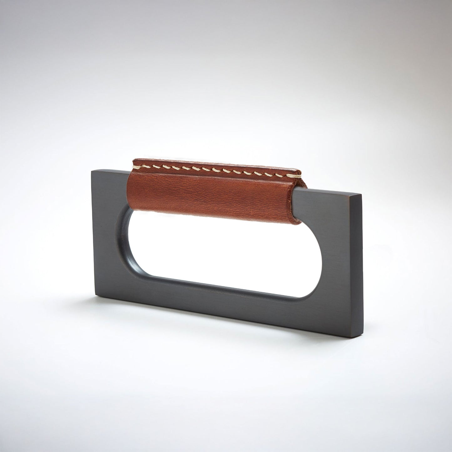 A modern rectangular metal cabinet pull with a central cutout, featuring a light brown leather strap stitched across the top, shown on a clean grey studio background.