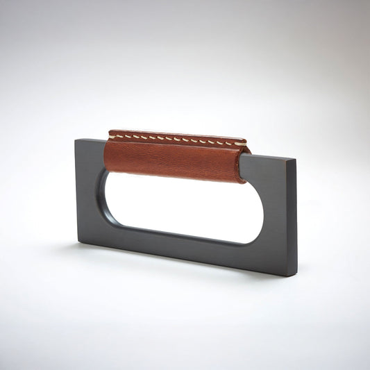 A modern rectangular metal cabinet pull with a central cutout, featuring a light brown leather strap stitched across the top, shown on a clean grey studio background.
