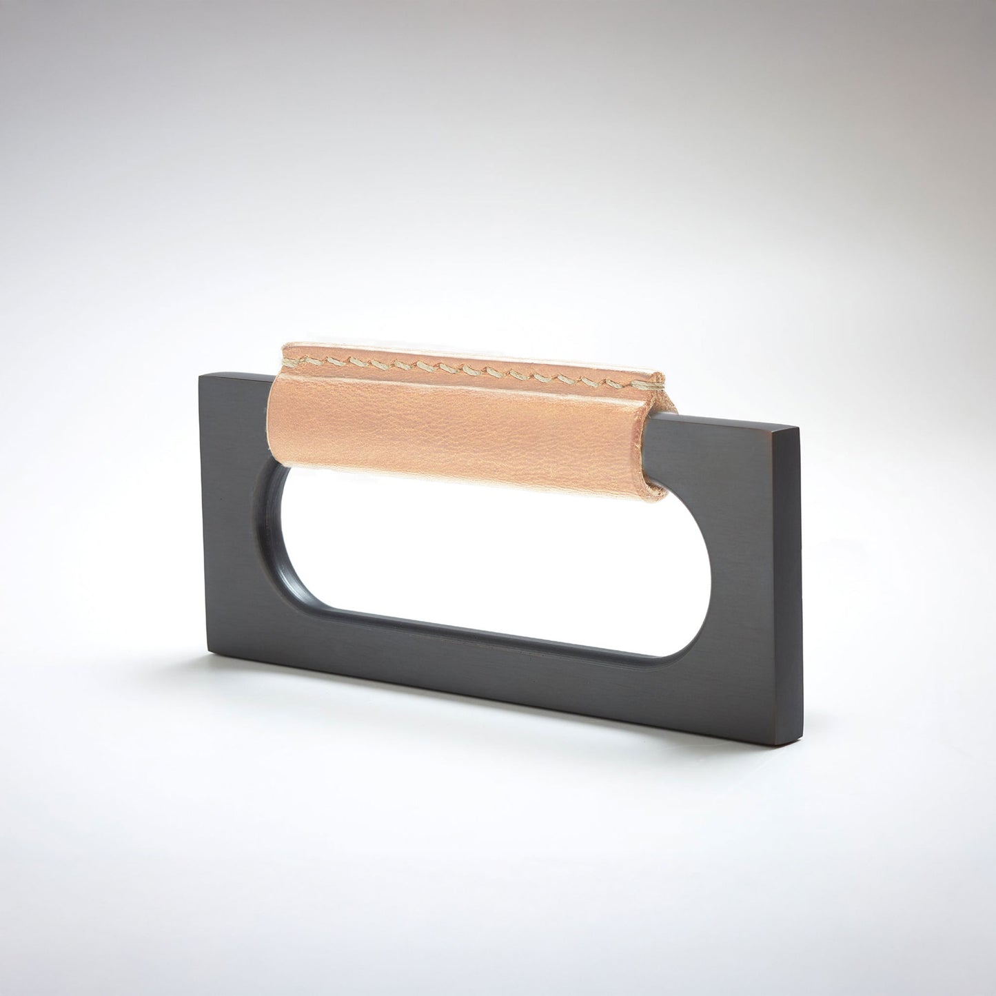 A modern rectangular metal cabinet pull with a central cutout, featuring a light tan leather strap stitched across the top, shown on a clean grey studio background.